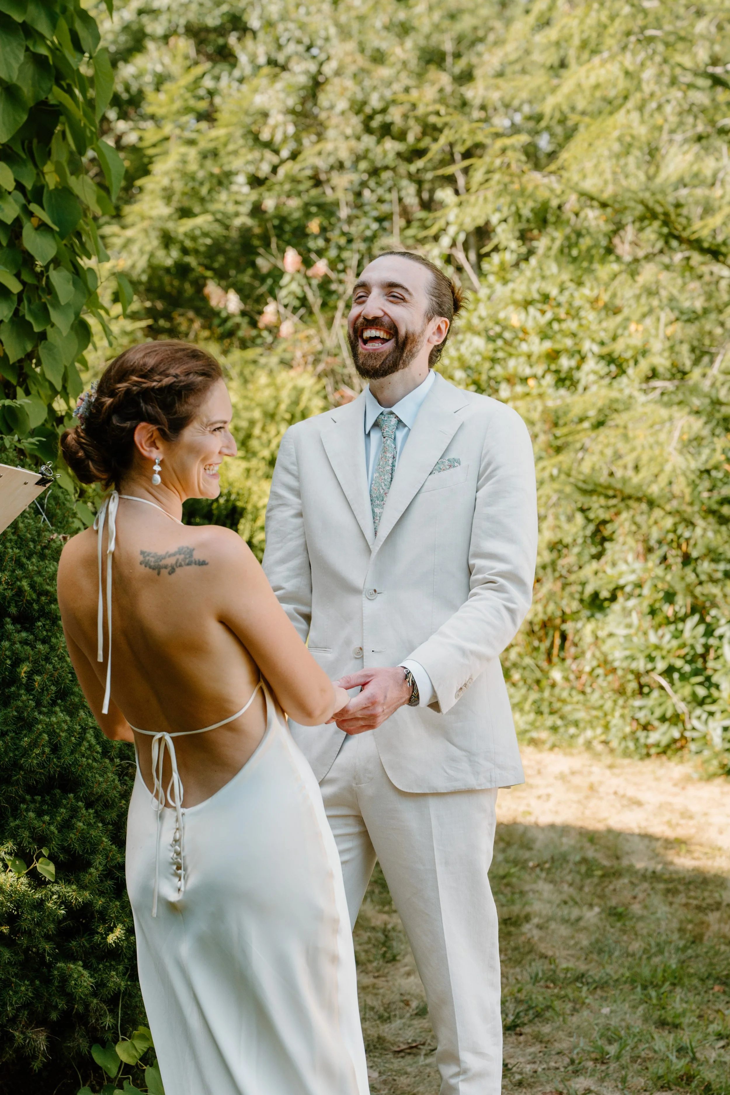 A couple is getting married outdoors, holding hands, and smiling at each other, with lush greenery in the background at a wedding in Bath, Maine. 