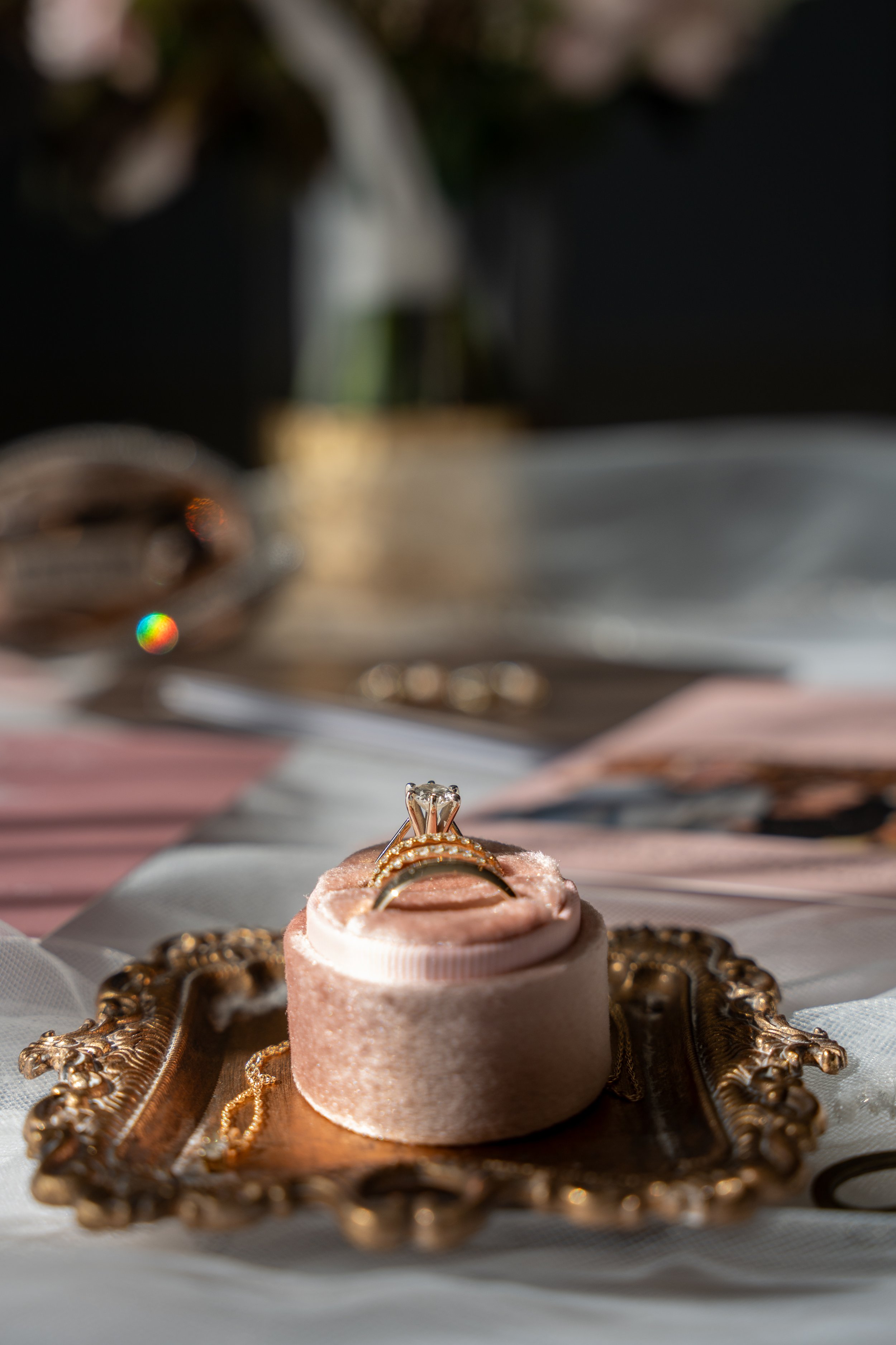 Close-up of a jewelry box with a diamond engagement ring and wedding band resting on top, placed on a decorative golden tray, with blurred background including a pink ribbon and photographs during a wedding at Red Jacket Resort in Cape Cod. 
