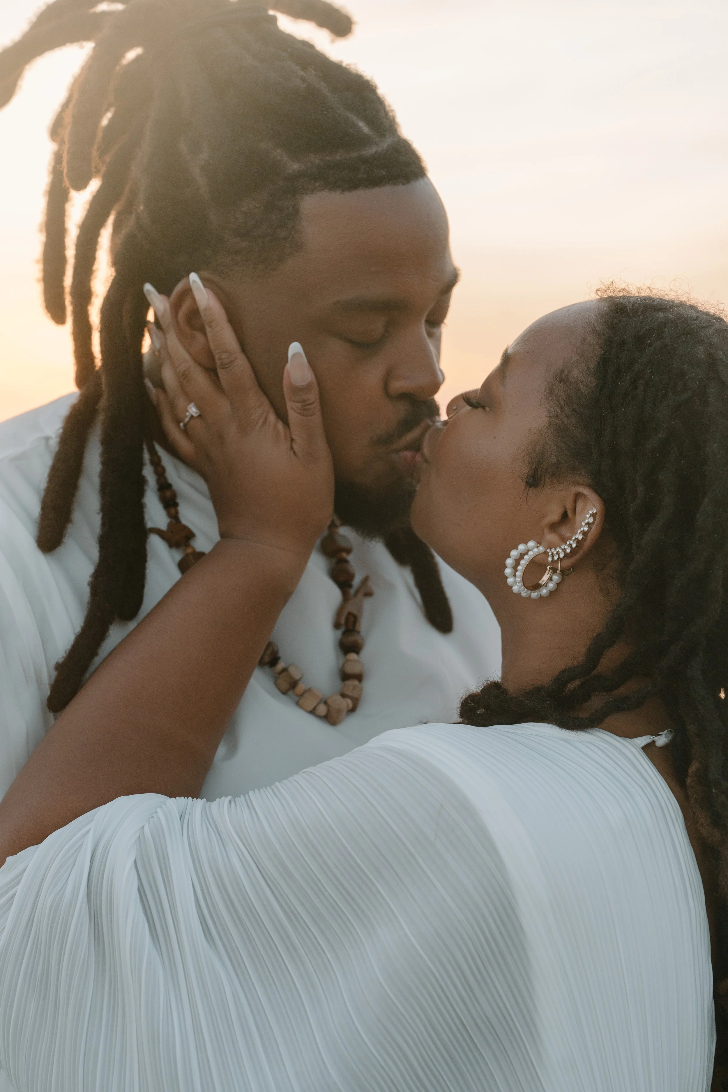 A couple sharing a kiss outdoors at sunset, with the man having dreadlocks and the woman wearing pearl earrings and a white dress at an elopement at Second Beach in Rhode Island. 