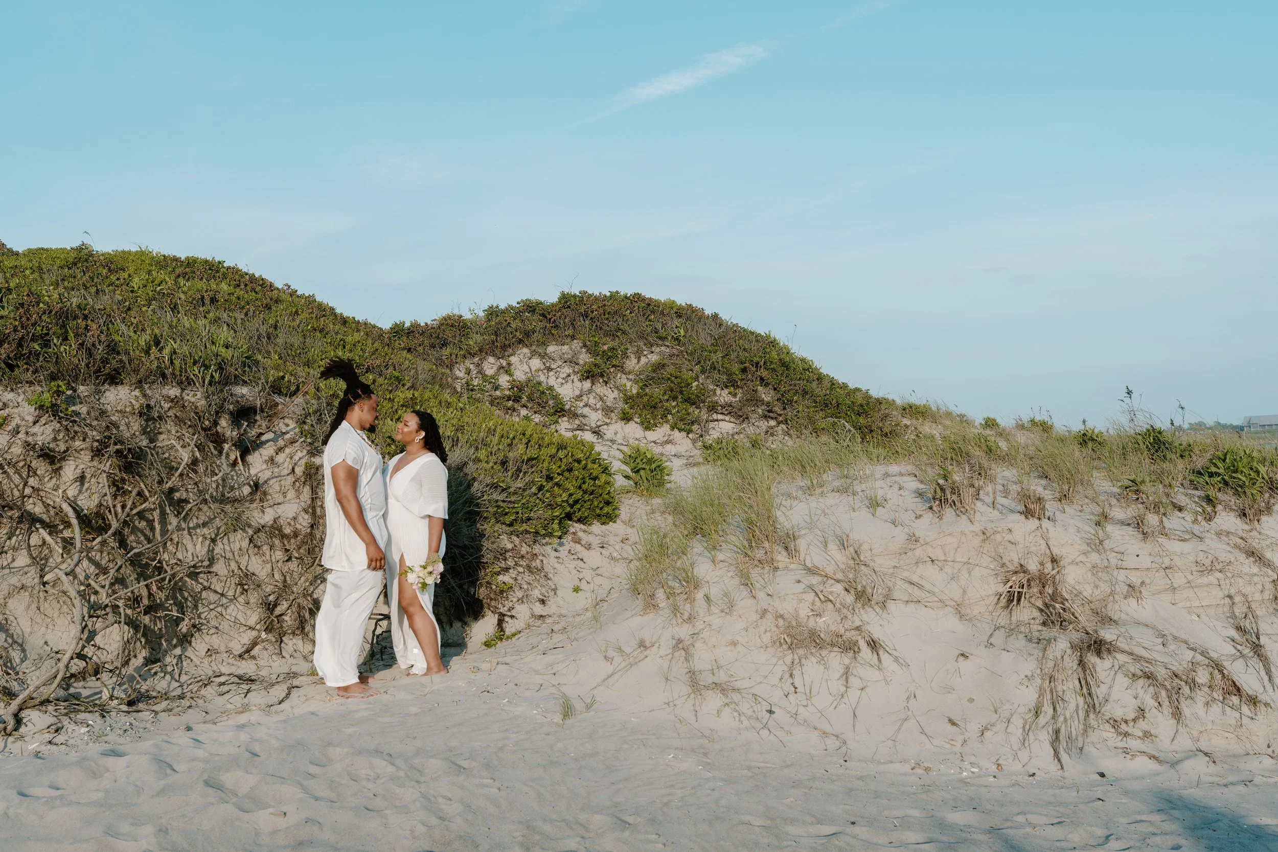 A couple dressed in white standing close together on a sandy beach near green dunes, with a clear blue sky overhead at an elopement at Second Beach in Rhode Island. 