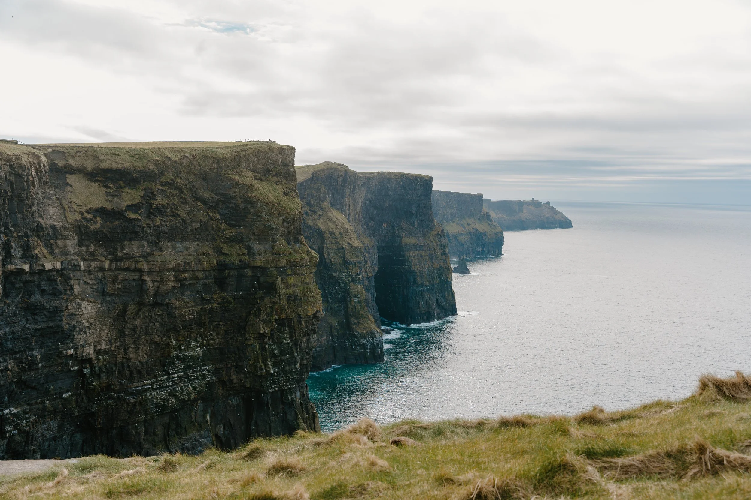 Cliffs stretching into the distance along a coastline, with grassy terrain in the foreground and ocean waves at the base of the cliffs under a cloudy sky for an engagement photo session in Galway, Ireland. 