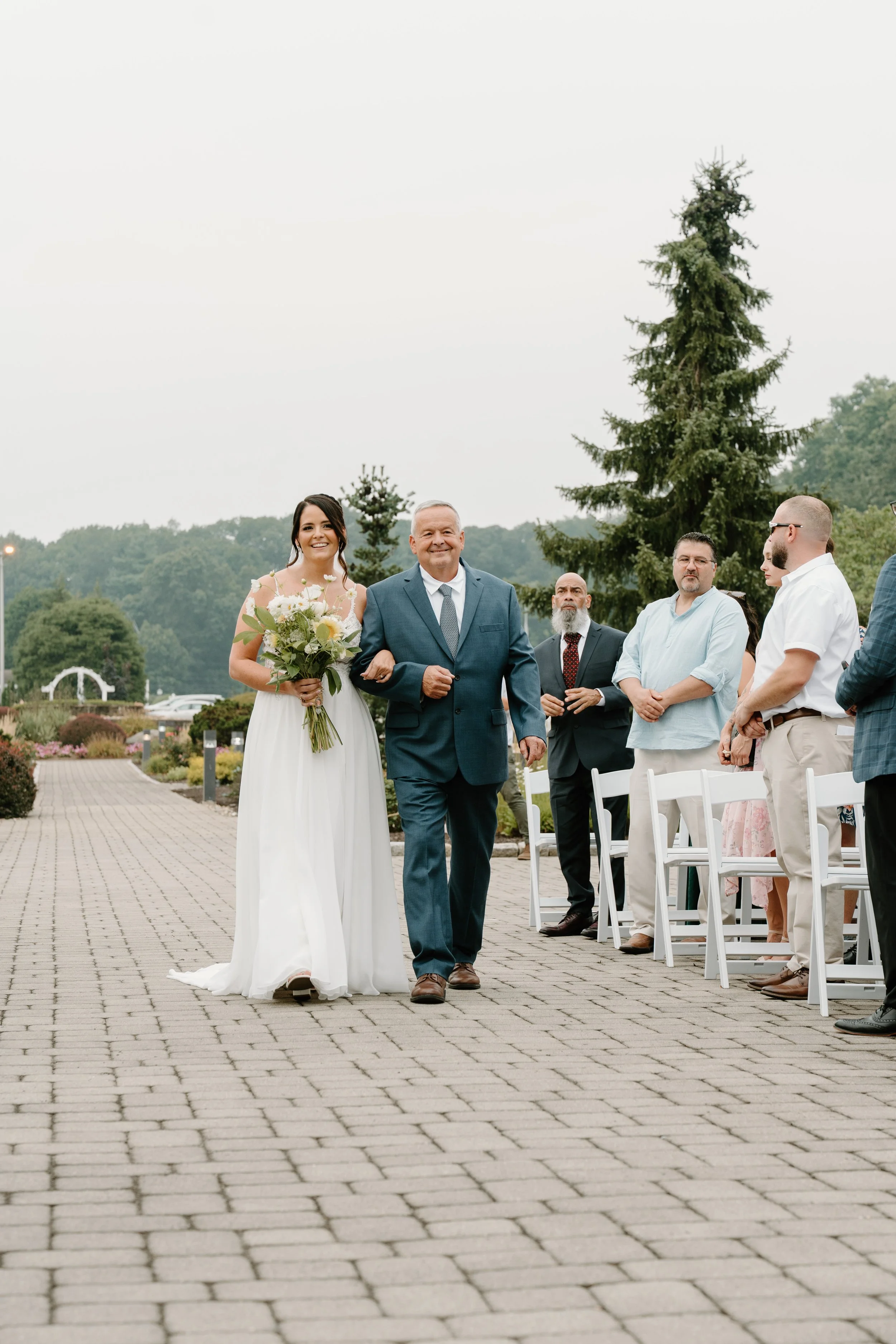 Bride walking down the aisle with her father at an outdoor wedding ceremony, surrounded by seated guests and scenic greenery during a wedding at the Aqua Turf Club in Connecticut. 
