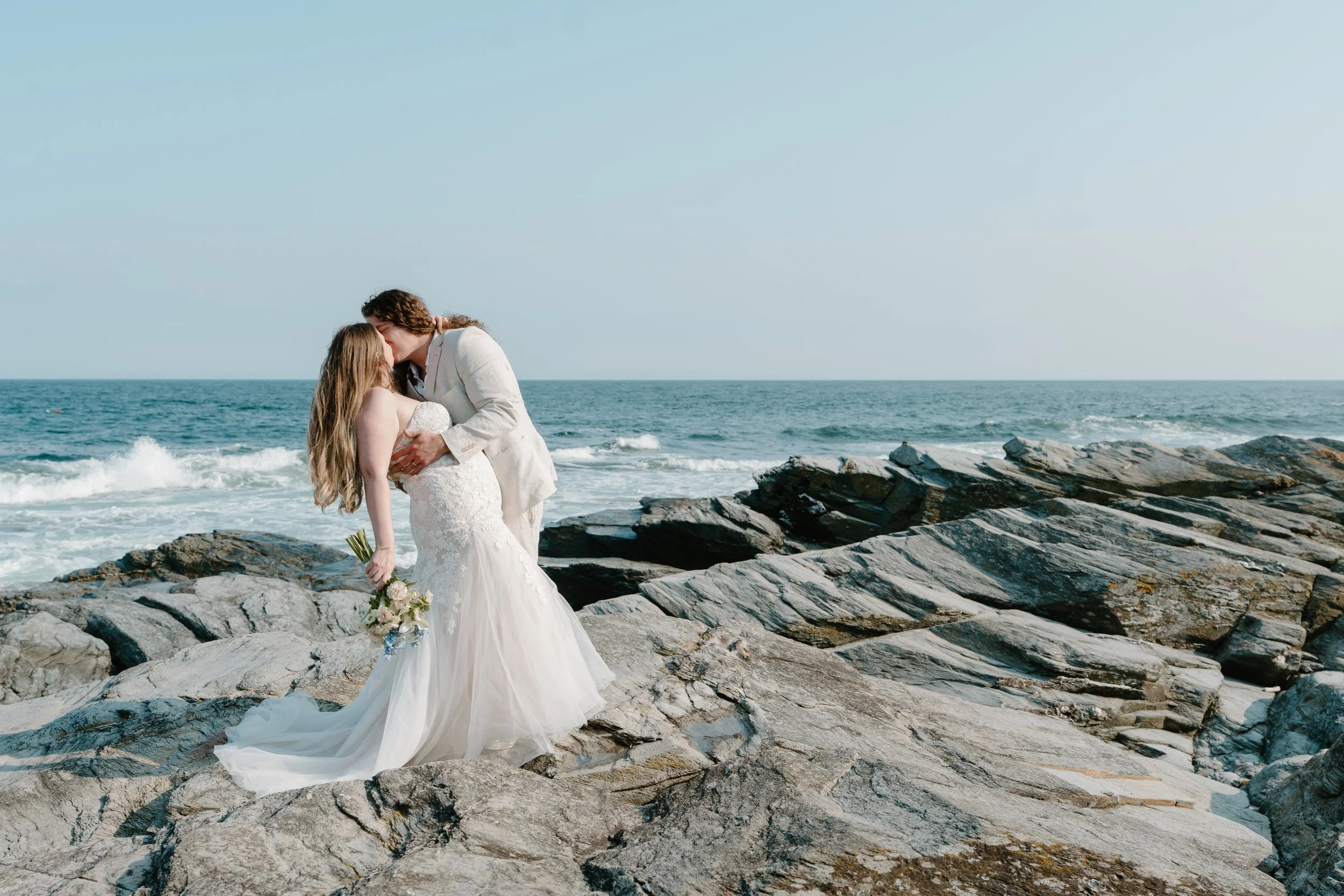 A bride and groom sharing a kiss on rocky beach with ocean waves in the background at an elopement at Beavertail State Park in Rhode Island. 