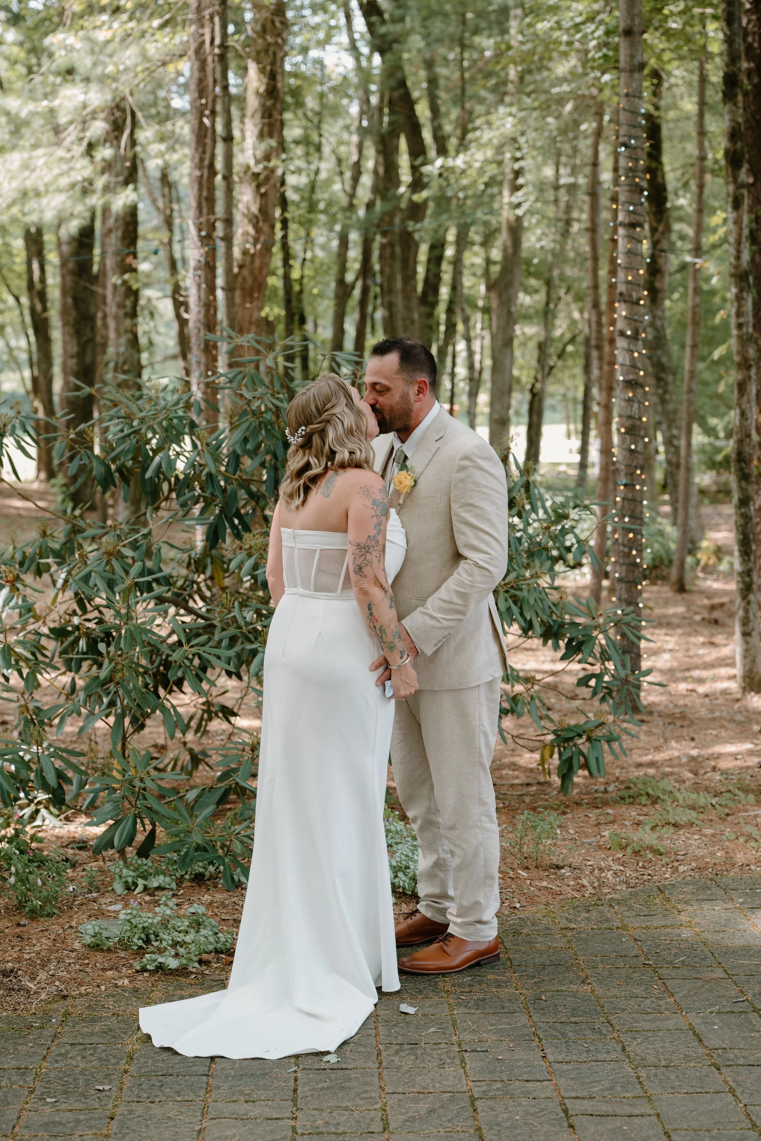 A couple in wedding attire sharing a kiss in a wooded outdoor setting with string lights around a tree.