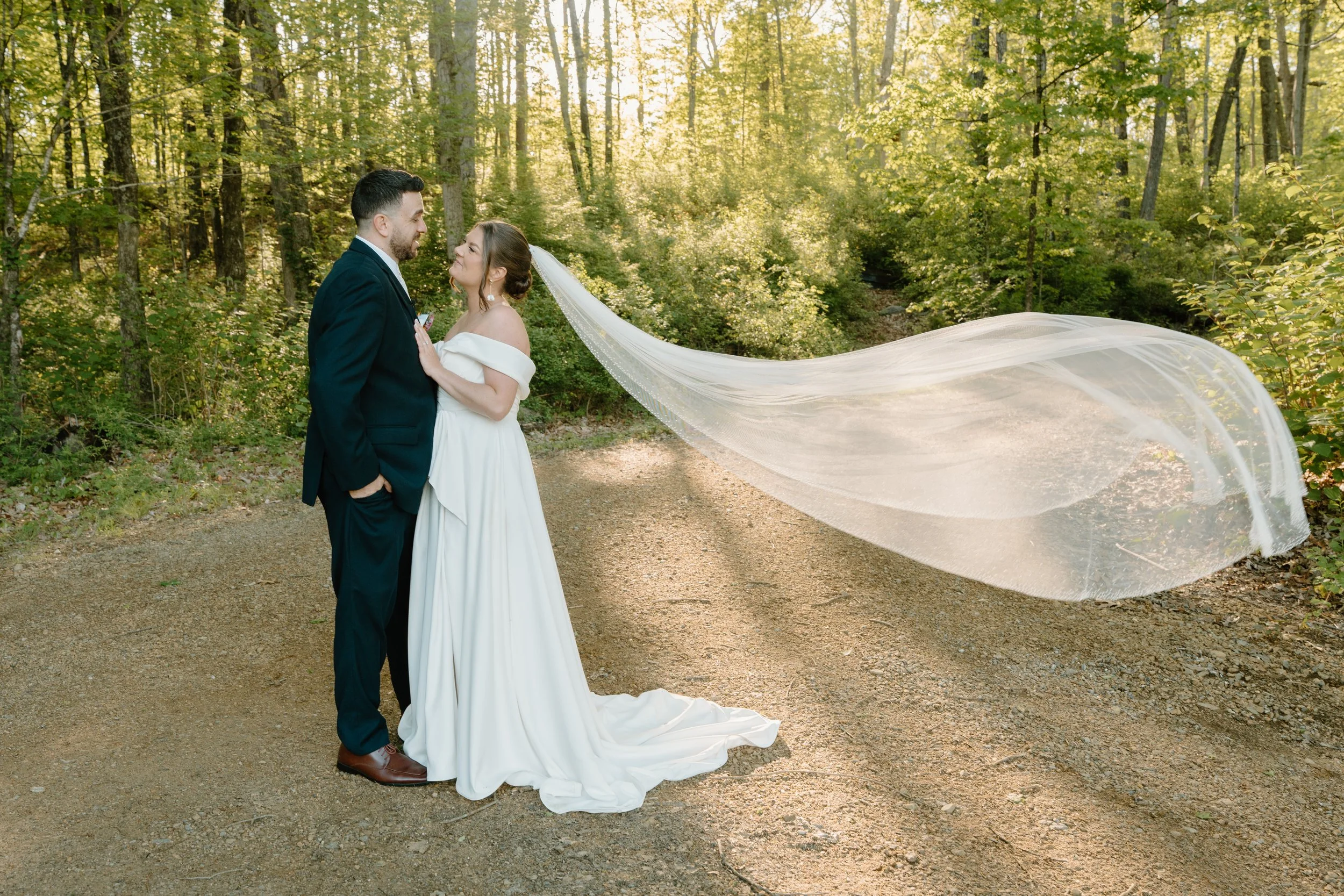 A bride and groom in wedding attire standing on a dirt path in a wooded area during daylight, with trees and sunlight in the background. The bride's veil is flowing behind her for a wedding at Holiday Hill Day Camp in Connecticut.