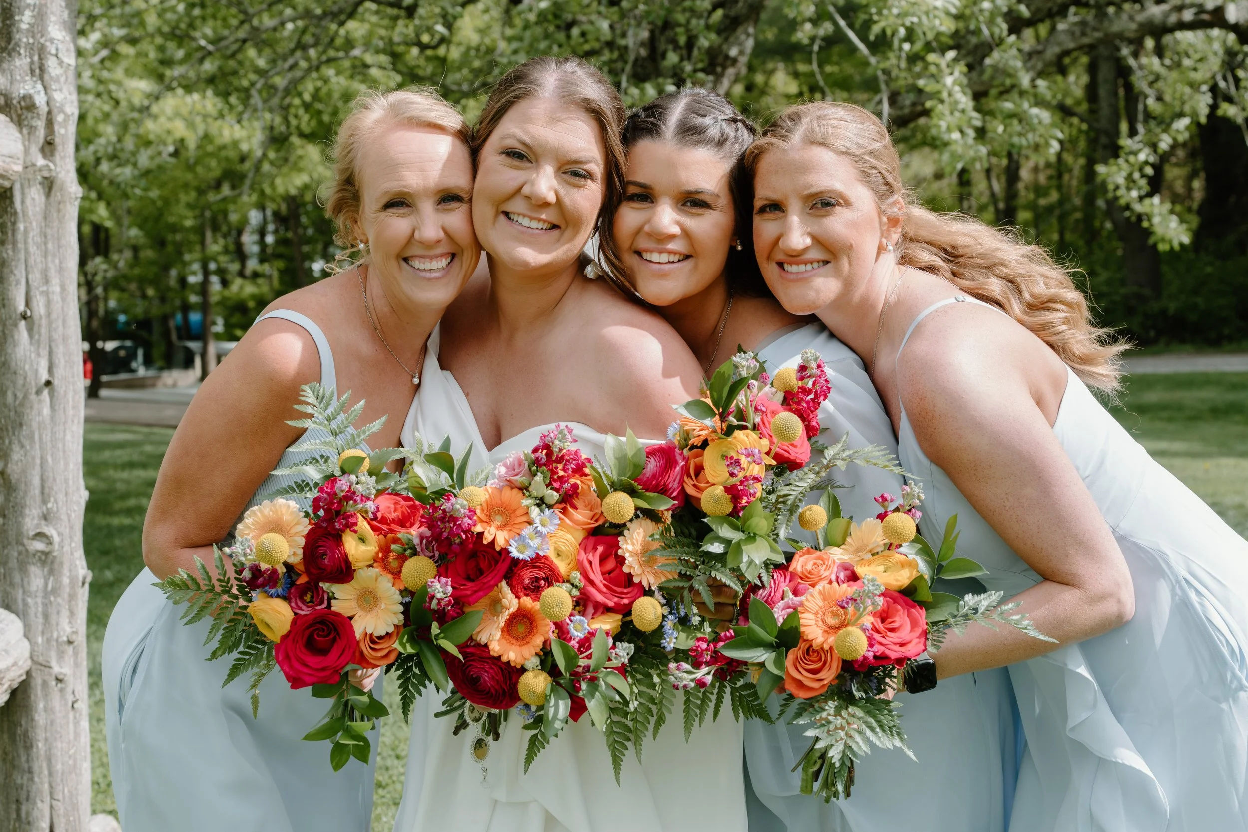 Four women in white dresses smiling and hugging, holding a large colorful wedding bouquet outdoors in a park with trees for a wedding at Holiday Hill Day Camp in Connecticut.