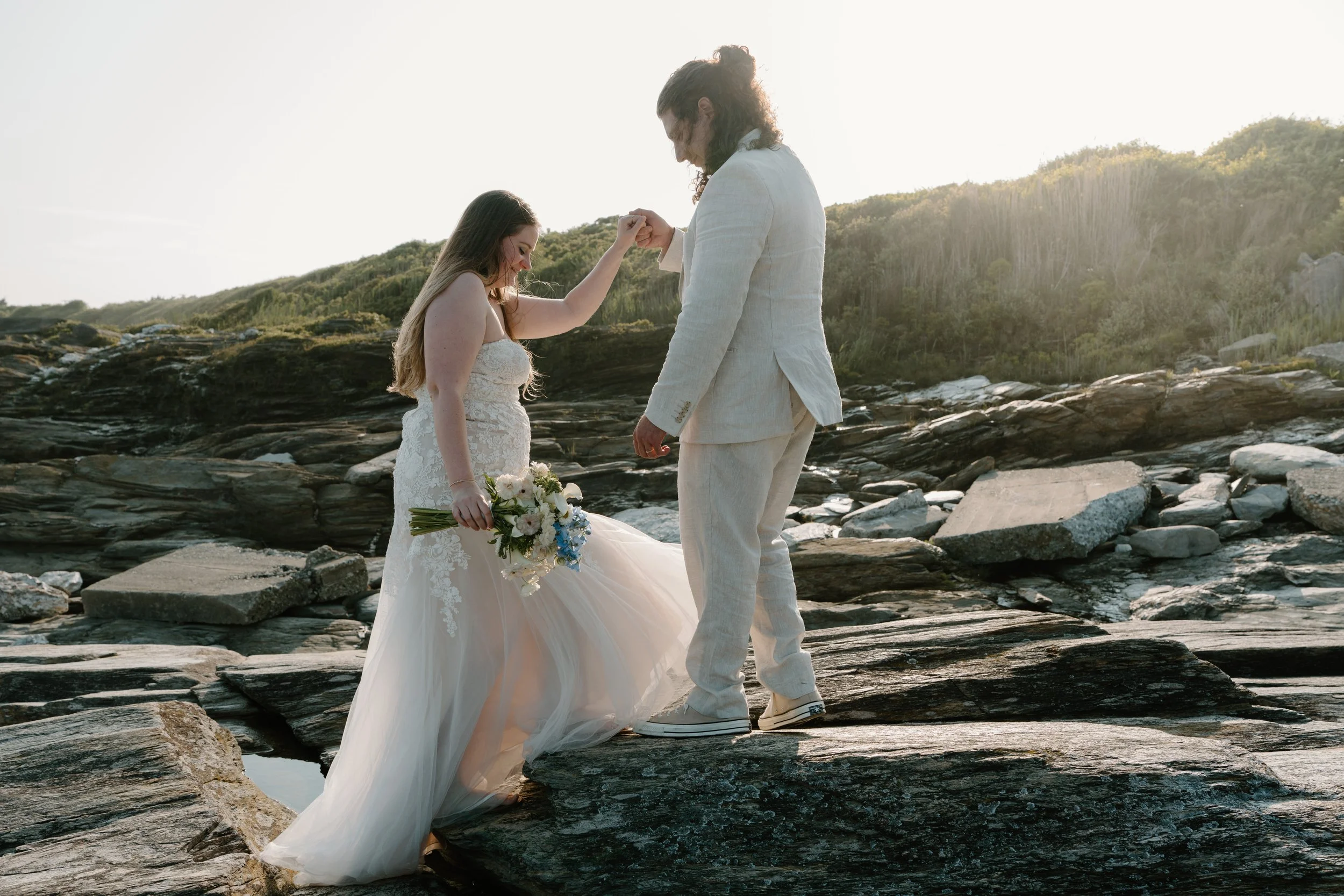 Bride and groom standing on rocks outdoors, holding hands, with the bride holding a bouquet, during sunset at an elopement at Beavertail State Park in Rhode Island. 