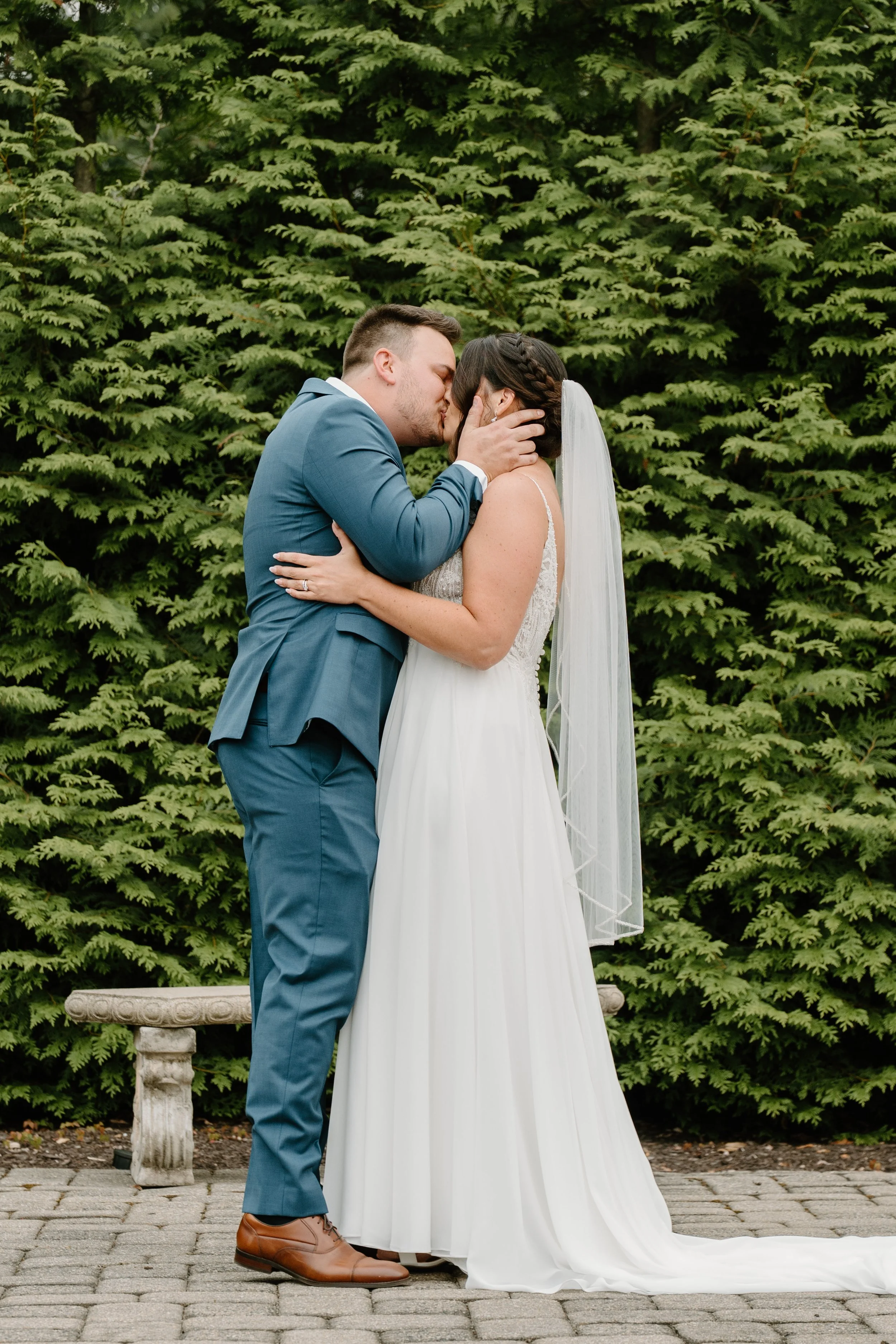 A bride and groom sharing a kiss outdoors on a paved area, with green foliage in the background. The bride wears a white wedding dress and veil, and the groom is in a blue suit during a wedding at the Aqua Turf Club in Connecticut. 