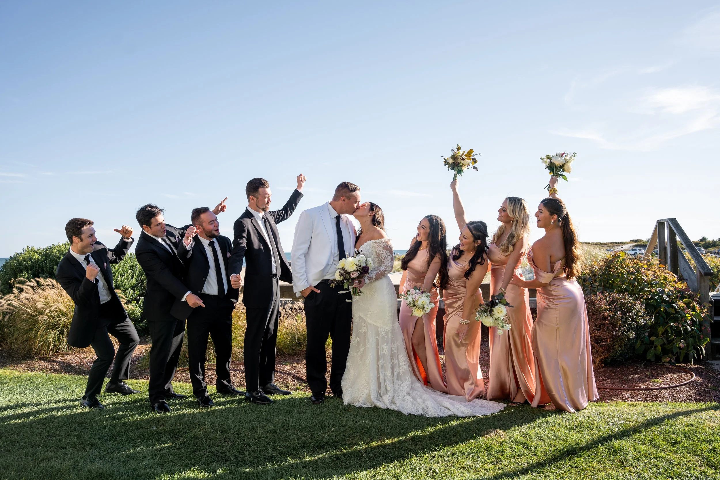 Bride and groom kissing outside, surrounded by wedding party, including bridesmaids and groomsmen celebrating with high-fives and holding bouquets during a wedding at Red Jacket Resort in Cape Cod. 