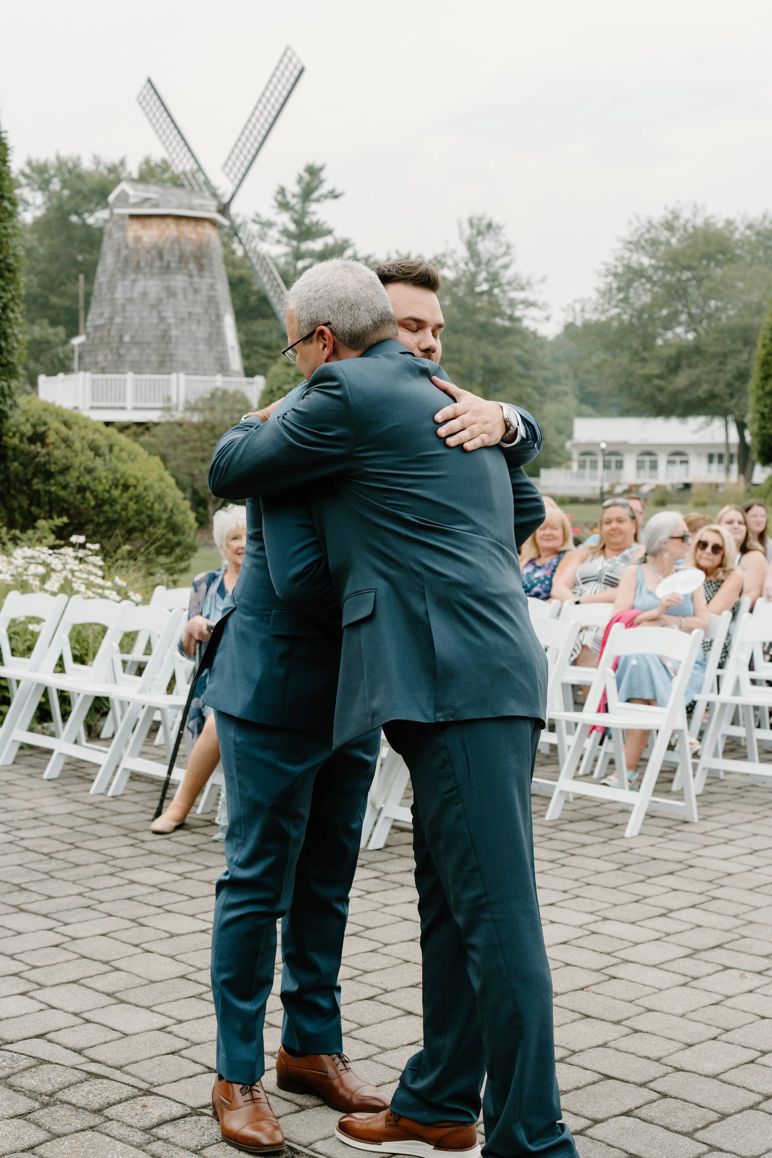 Two men in suits hugging at an outdoor event with white chairs, a small crowd of seated guests, a white building, and a windmill in the background during a wedding at the Aqua Turf Club in Connecticut. 