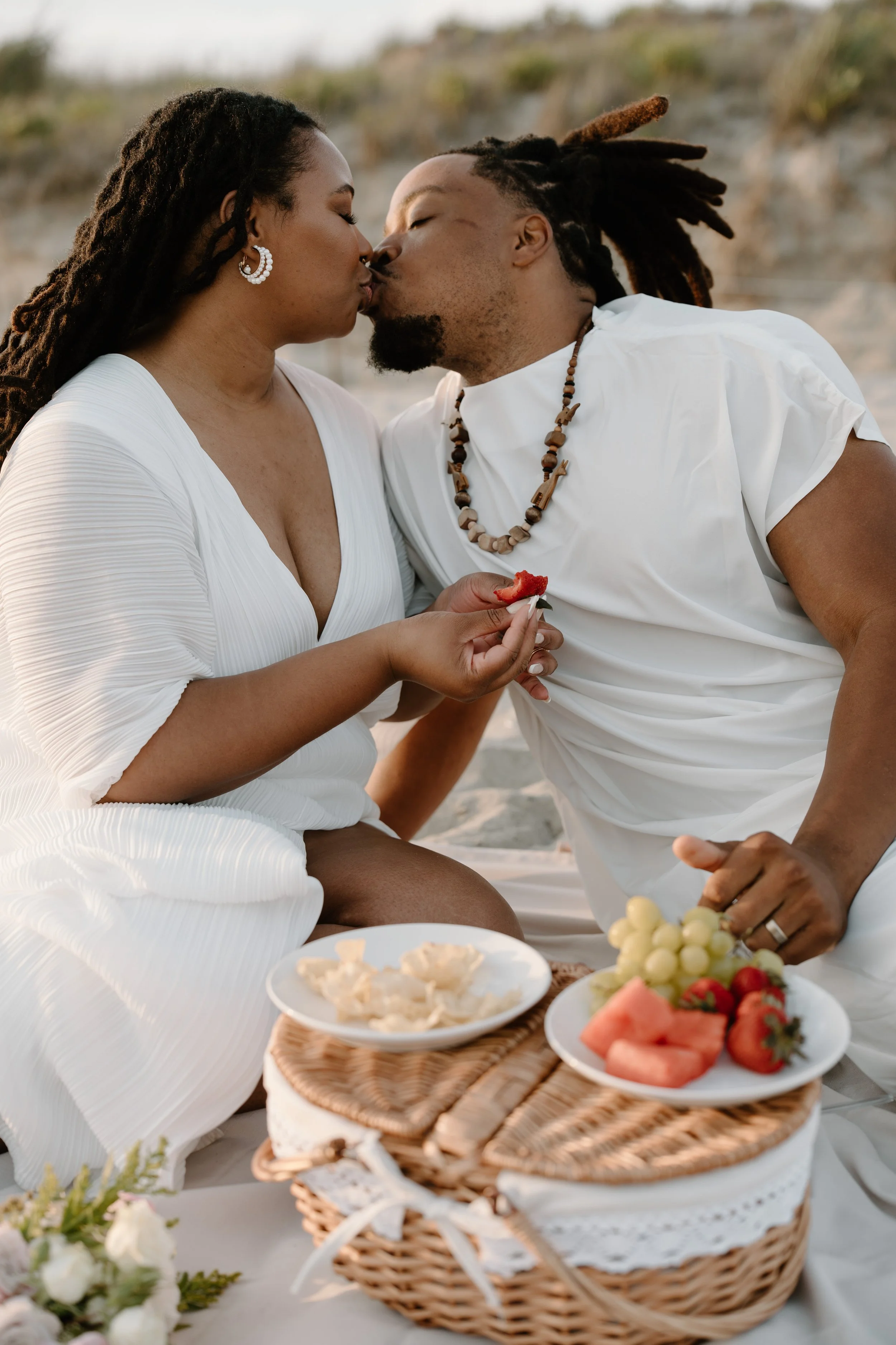 A couple sharing a kiss during a beach picnic, sitting on a blanket with baskets of food and plates of fresh fruit at an elopement at Second Beach in Rhode Island. 