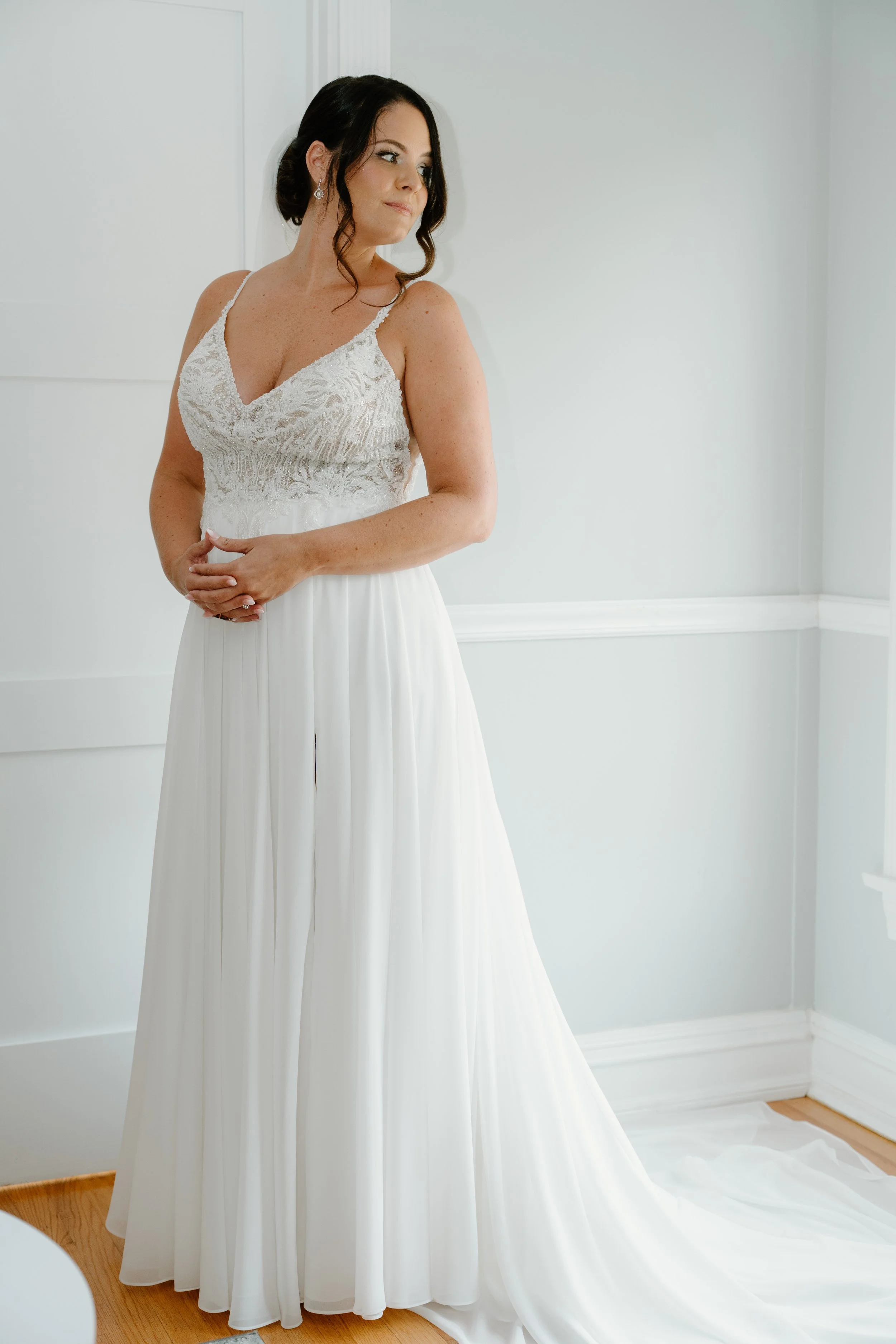 A woman in a white wedding dress, standing indoors near a corner, with dark hair styled in loose waves, looking to her left during a wedding at the Aqua Turf Club in Connecticut. 