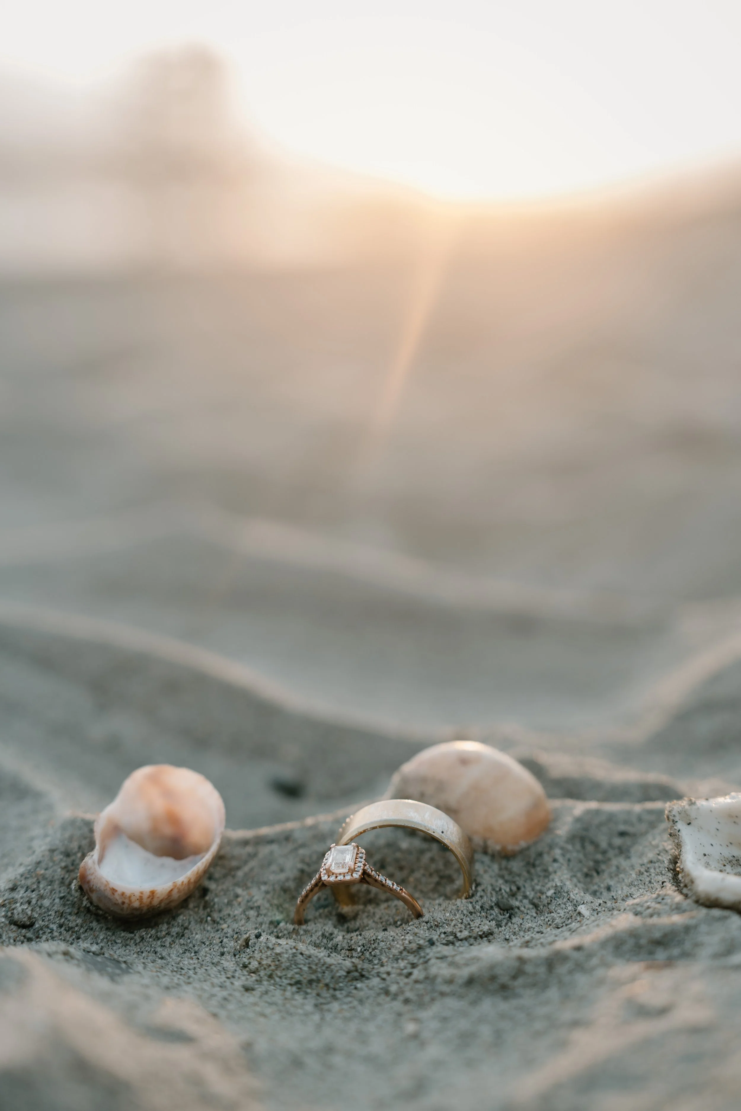 A close-up of a couple of rings, one with a crushed square diamond, set on sandy beach among seashells at sunset at an elopement at Second Beach in Rhode Island. 