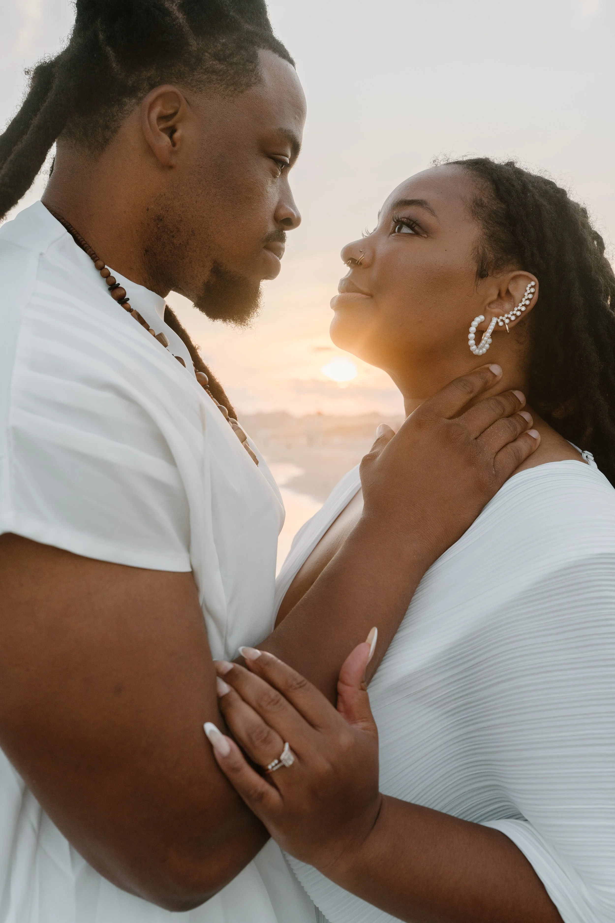 A couple gazes into each other's eyes during sunset, with the man gently holding the woman's neck and the woman embracing the man's arm. They wear white clothing and jewelry at an elopement at Second Beach in Rhode Island. 