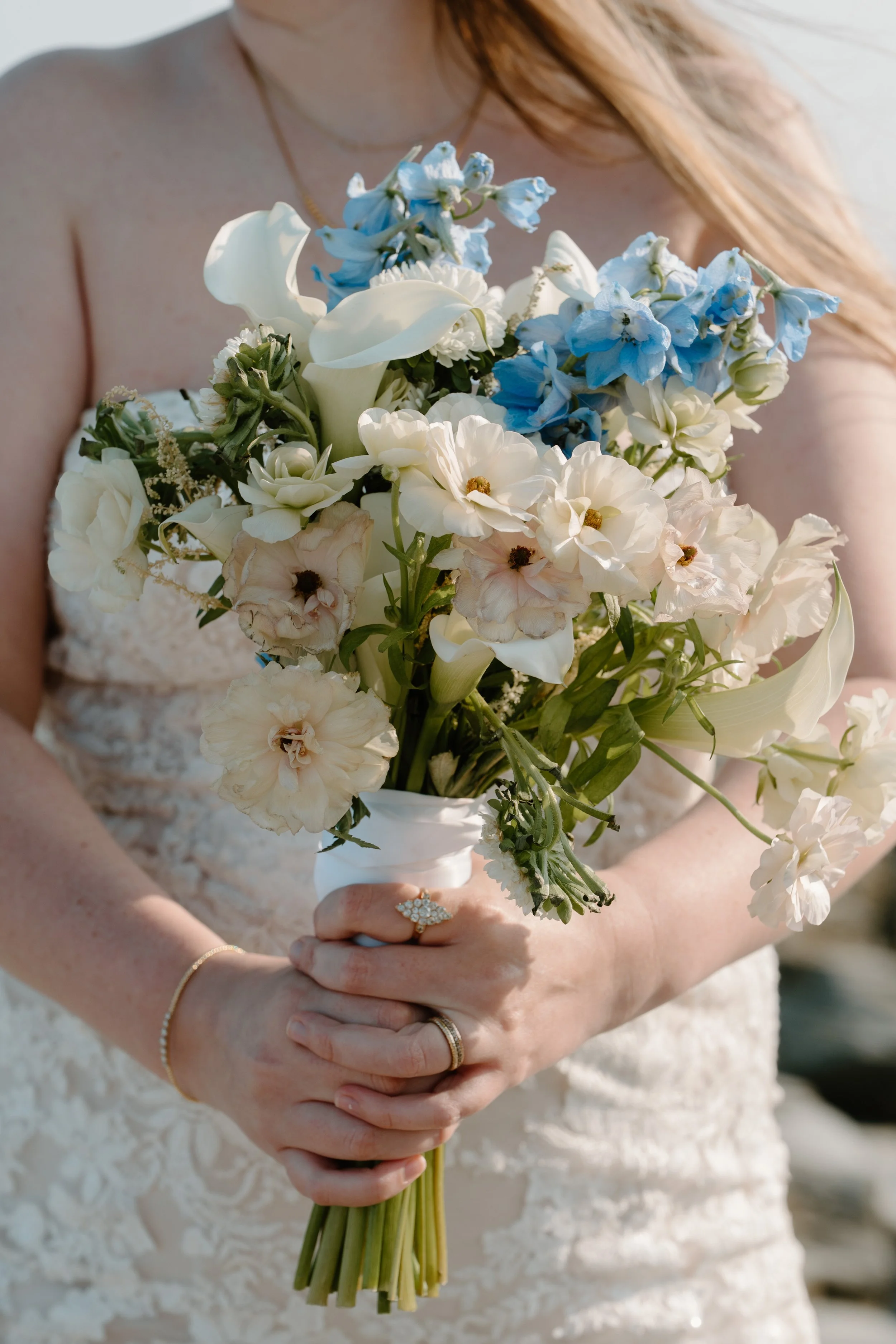 A woman wearing a wedding dress holding a bouquet of white, cream, and blue flowers at an elopement at Beavertail State Park in Rhode Island. 