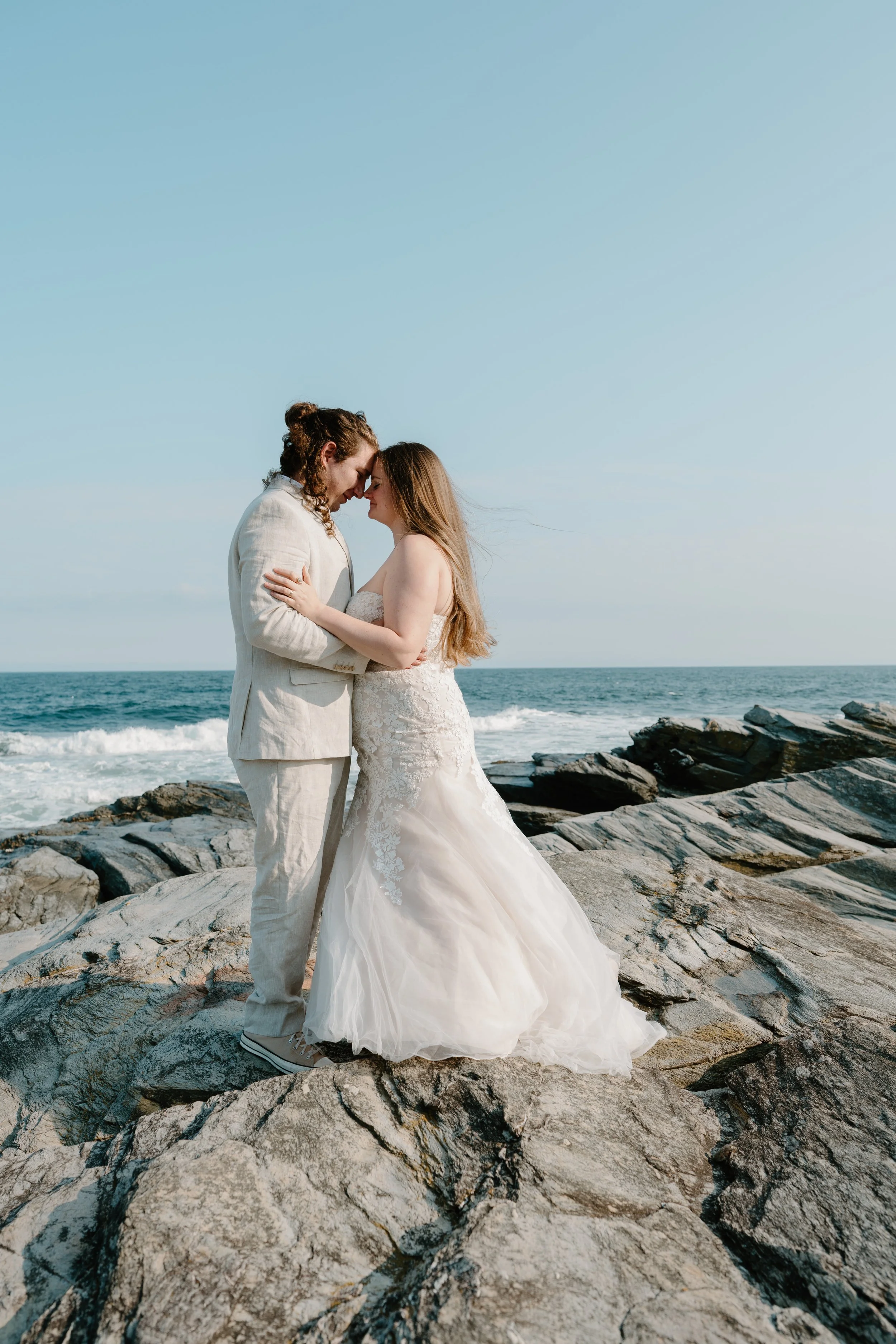 A couple in wedding attire embracing on rocks by the ocean with clear skies at an elopement at Beavertail State Park in Rhode Island. 