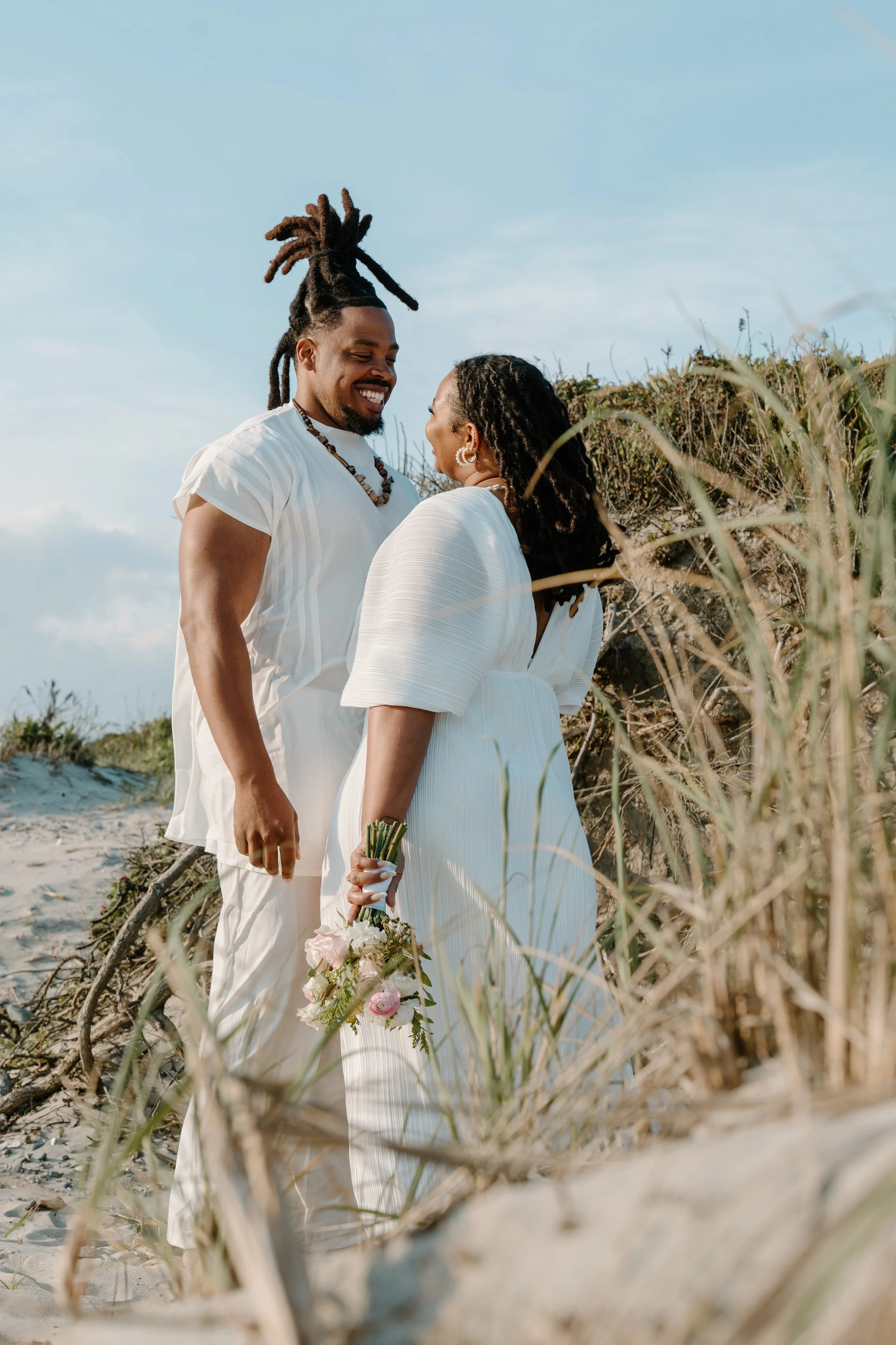 A couple dressed in white standing on a beach dune, smiling and looking at each other. The woman holds a bouquet of flowers at an elopement at Second Beach in Rhode Island. 