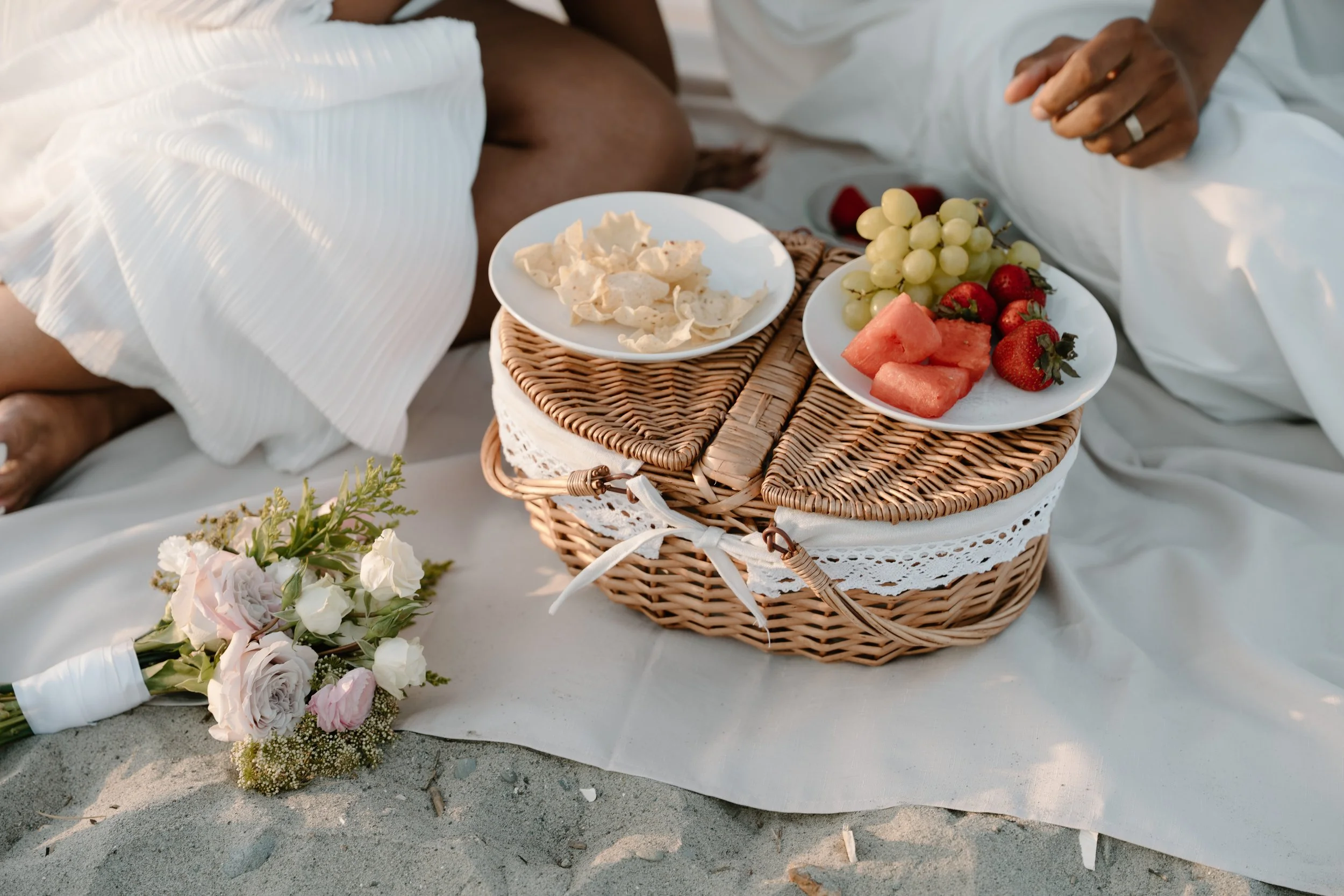 A picnic basket with plates of fresh fruit, crackers, and strawberries on a white sheet at the beach, with a bouquet of flowers nearby at an elopement at Second Beach in Rhode Island. 