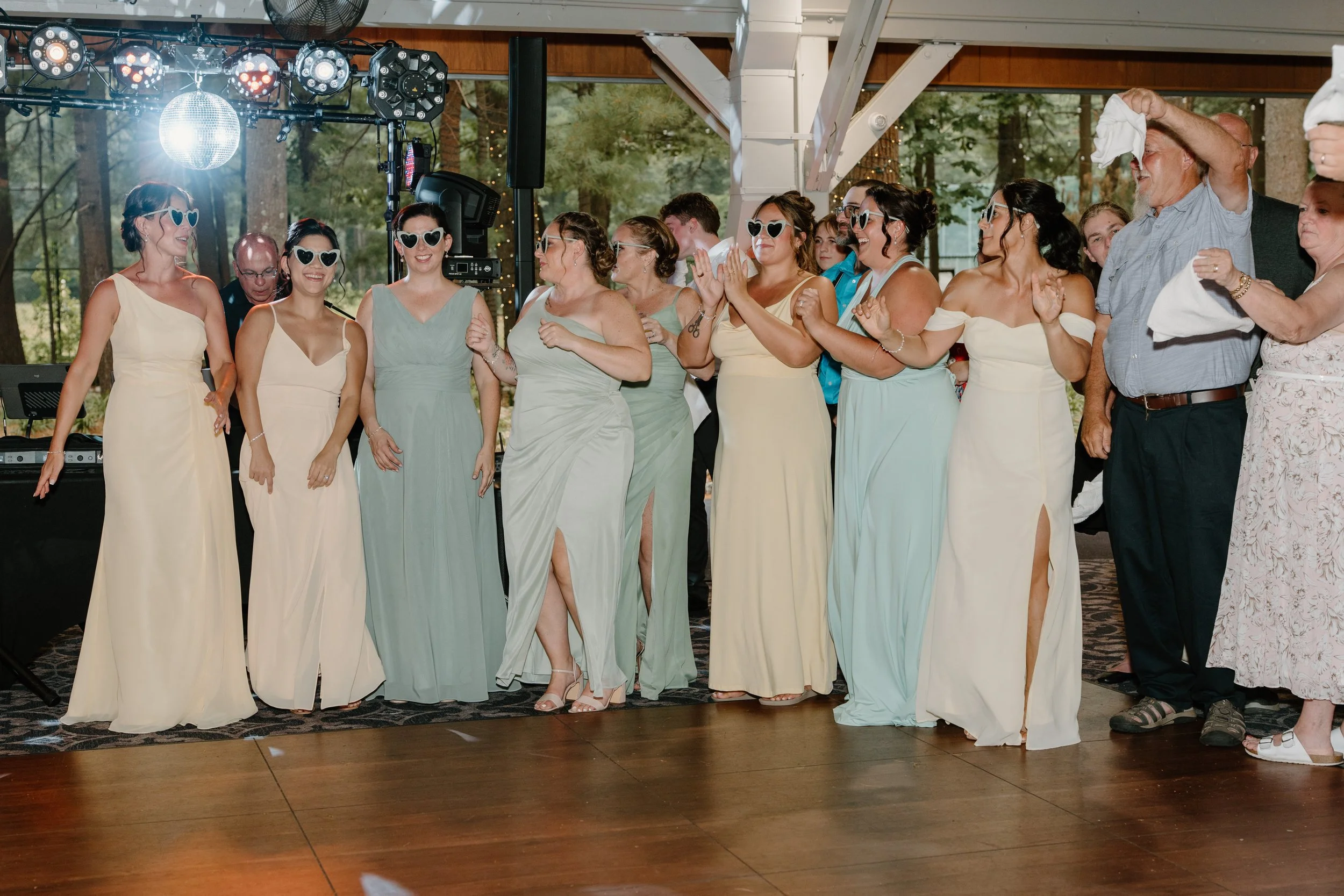 Group of women and men at a wedding reception, wearing pastel dresses, with some women wearing heart-shaped sunglasses, standing on a dance floor, smiling and clapping for a wedding at Tunxis Country Club in Connecticut. 