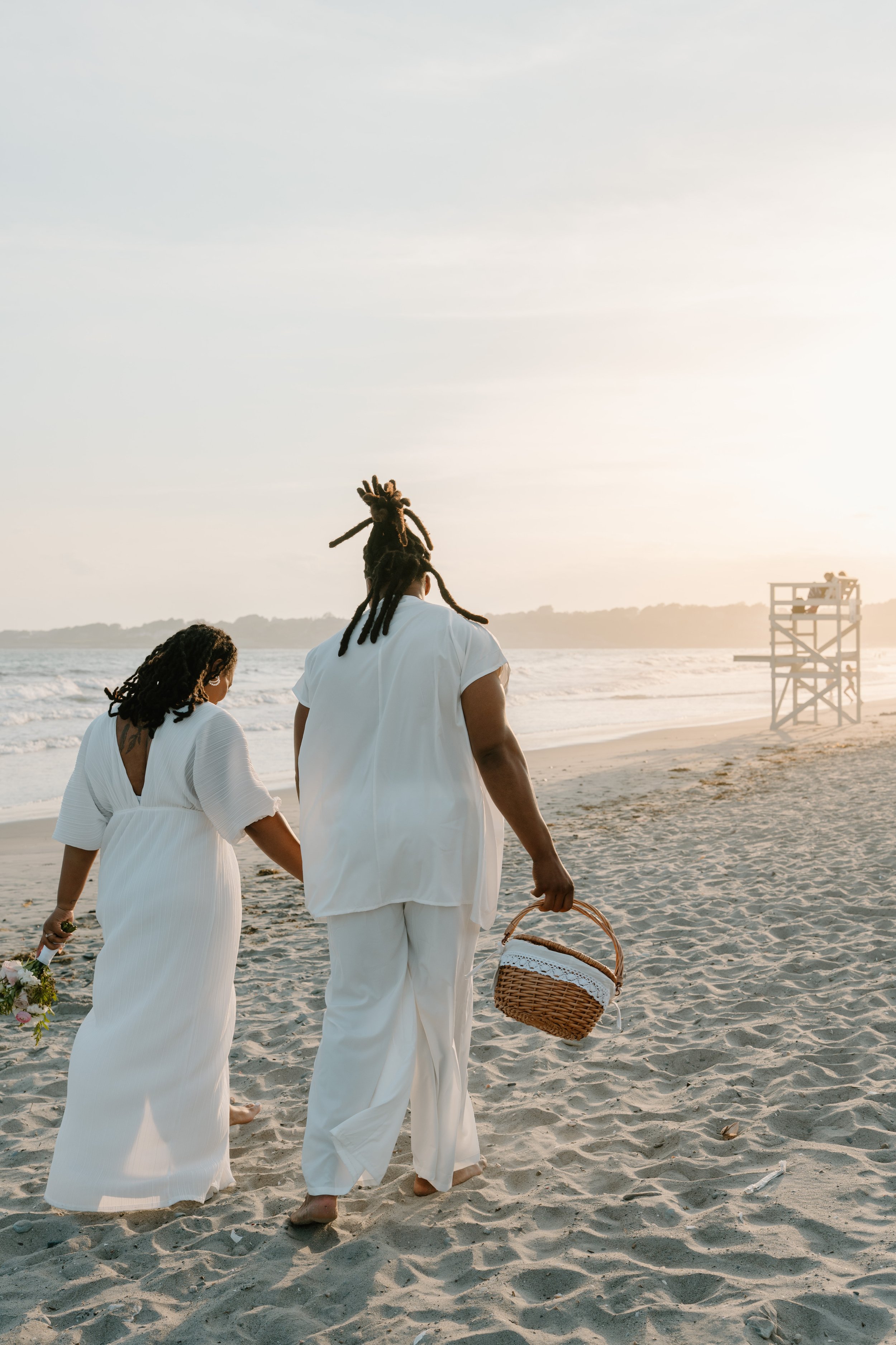 A couple walking along the beach holding hands during sunset, with the man carrying a picnic basket and the woman holding a bouquet of flowers at an elopement at Second Beach in Rhode Island. 
