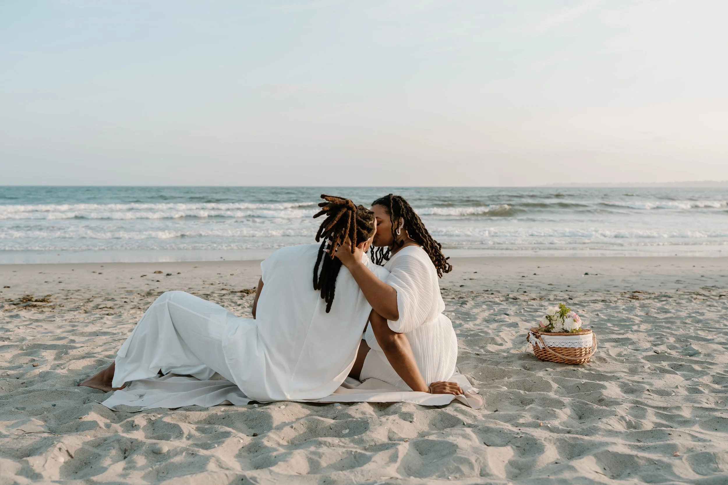 A couple dressed in white sharing a kiss on a sandy beach with the ocean in the background, a basket with flowers nearby at an elopement at Second Beach in Rhode Island. 