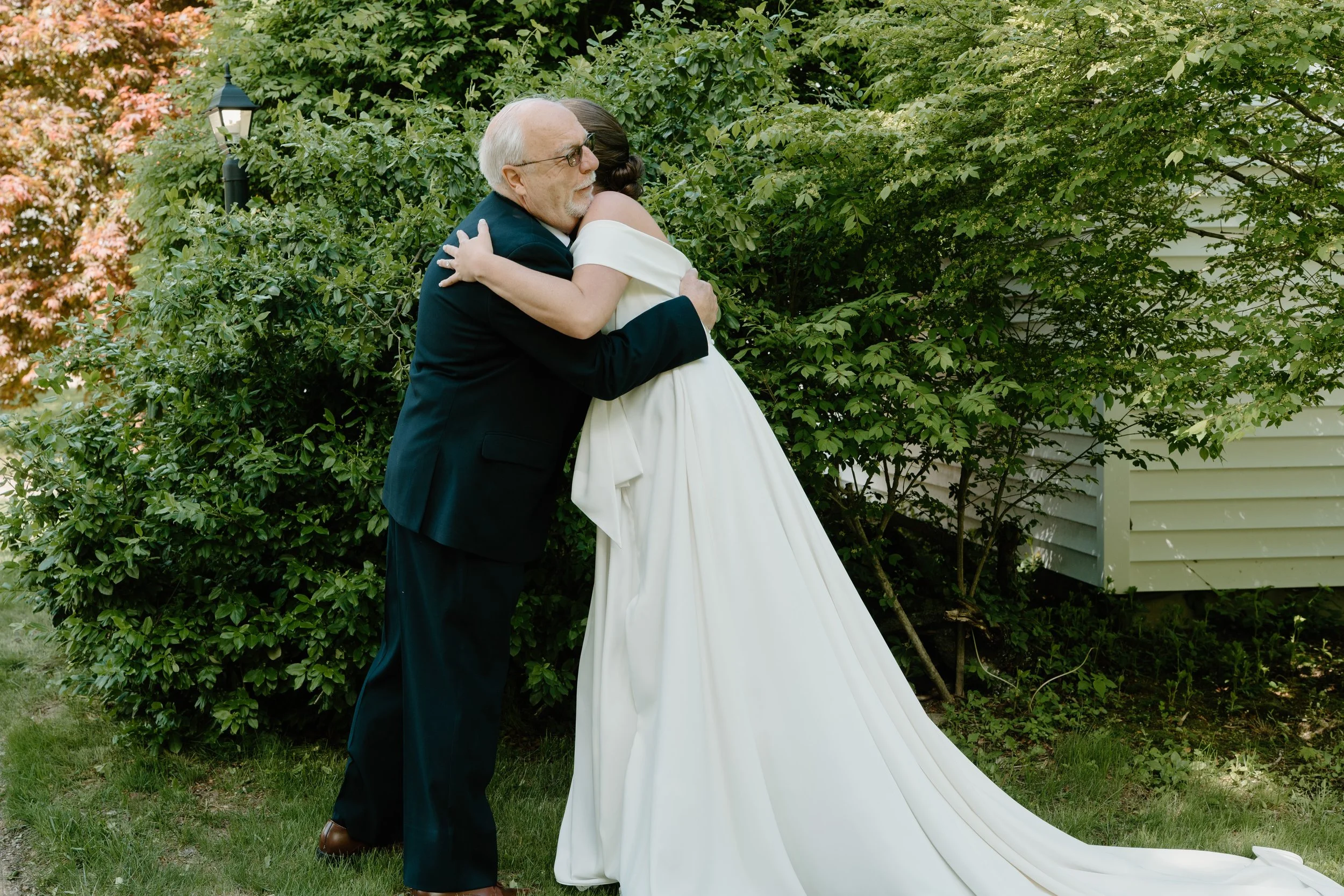 An bride and father first look in a black suit and a woman in a white wedding dress hugging outdoors in front of green bushes for a wedding at Holiday Hill Day Camp in Connecticut.
