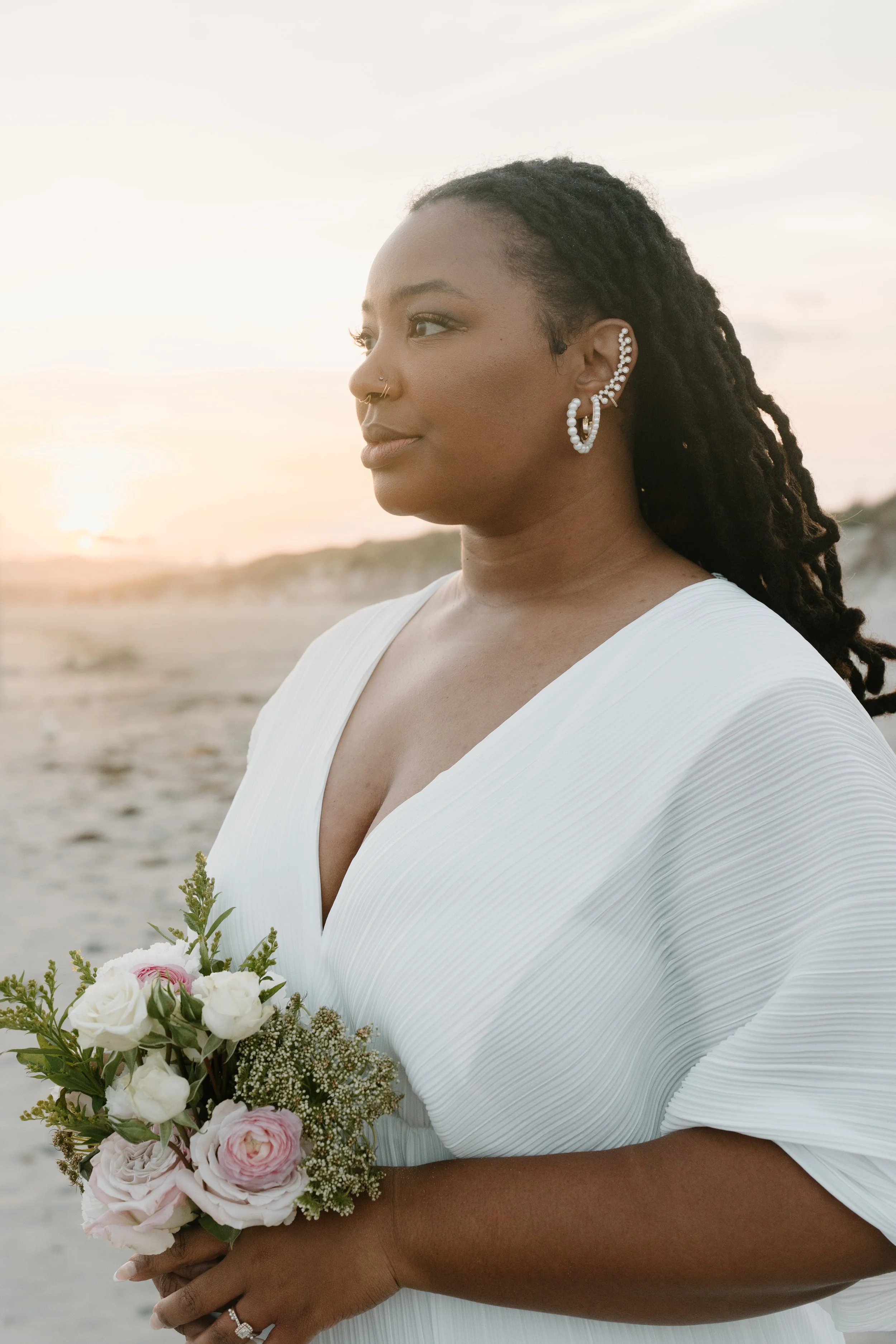 A woman wearing a white dress with pleats, holding a bouquet of pink and white flowers, standing on a beach at sunset, with her side profile visible at an elopement at Second Beach in Rhode Island. 