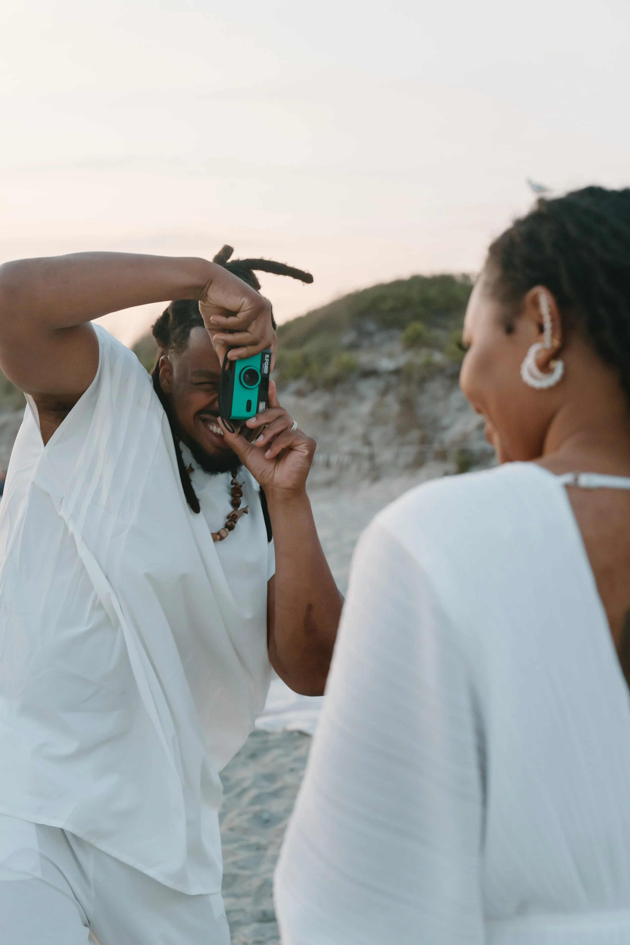 A man taking a picture with a camera, smiling, while a woman faces away watching on a beach at sunset at an elopement at Second Beach in Rhode Island. 