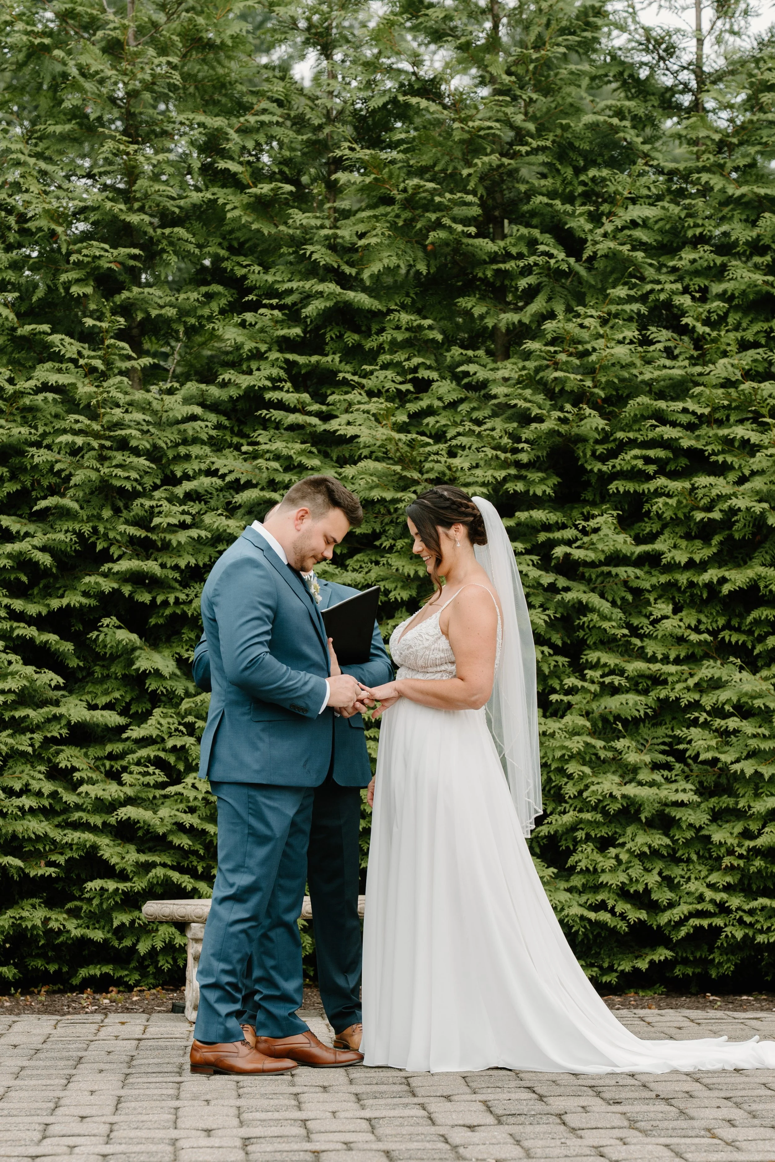 A couple gets married outdoors with a green hedge background, holding hands and smiling at each other, during their wedding ceremony during a wedding at the Aqua Turf Club in Connecticut. 