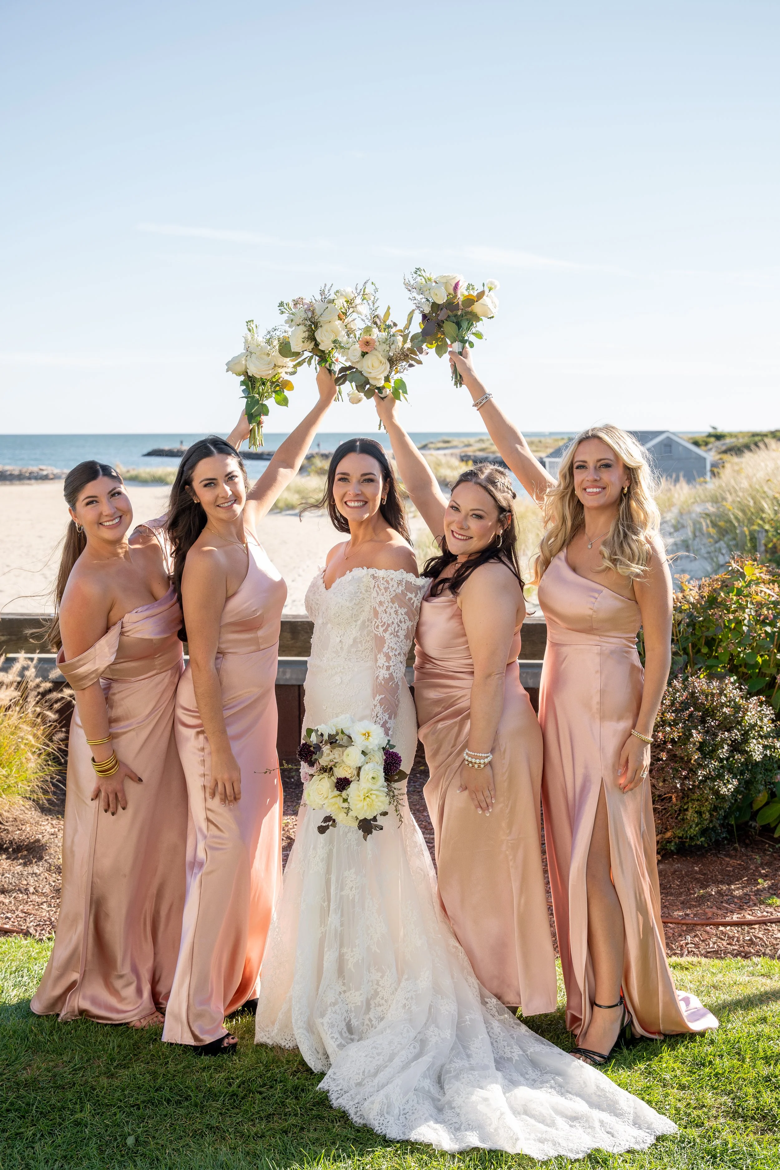 Group of five women celebrating on a beach, with the bride in the center holding a bouquet, and bridesmaids in matching blush dresses holding up flower arrangements during a wedding at Red Jacket Resort in Cape Cod. 