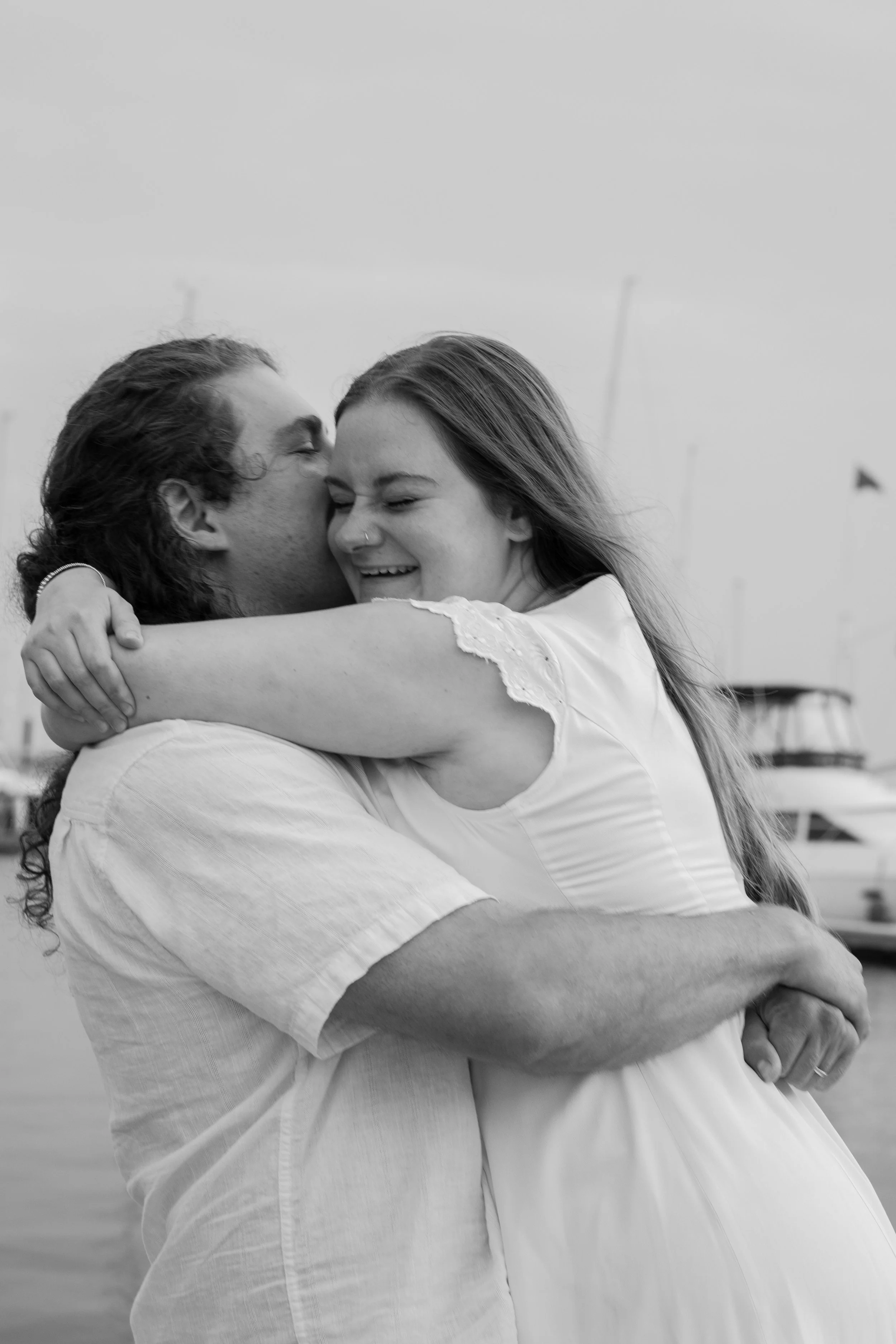 A black-and-white photo of a couple embracing on a boat dock, with the man kissing the woman on the forehead and the woman smiling with closed eyes, arms wrapped around each other at an elopement at Beavertail State Park in Rhode Island. 