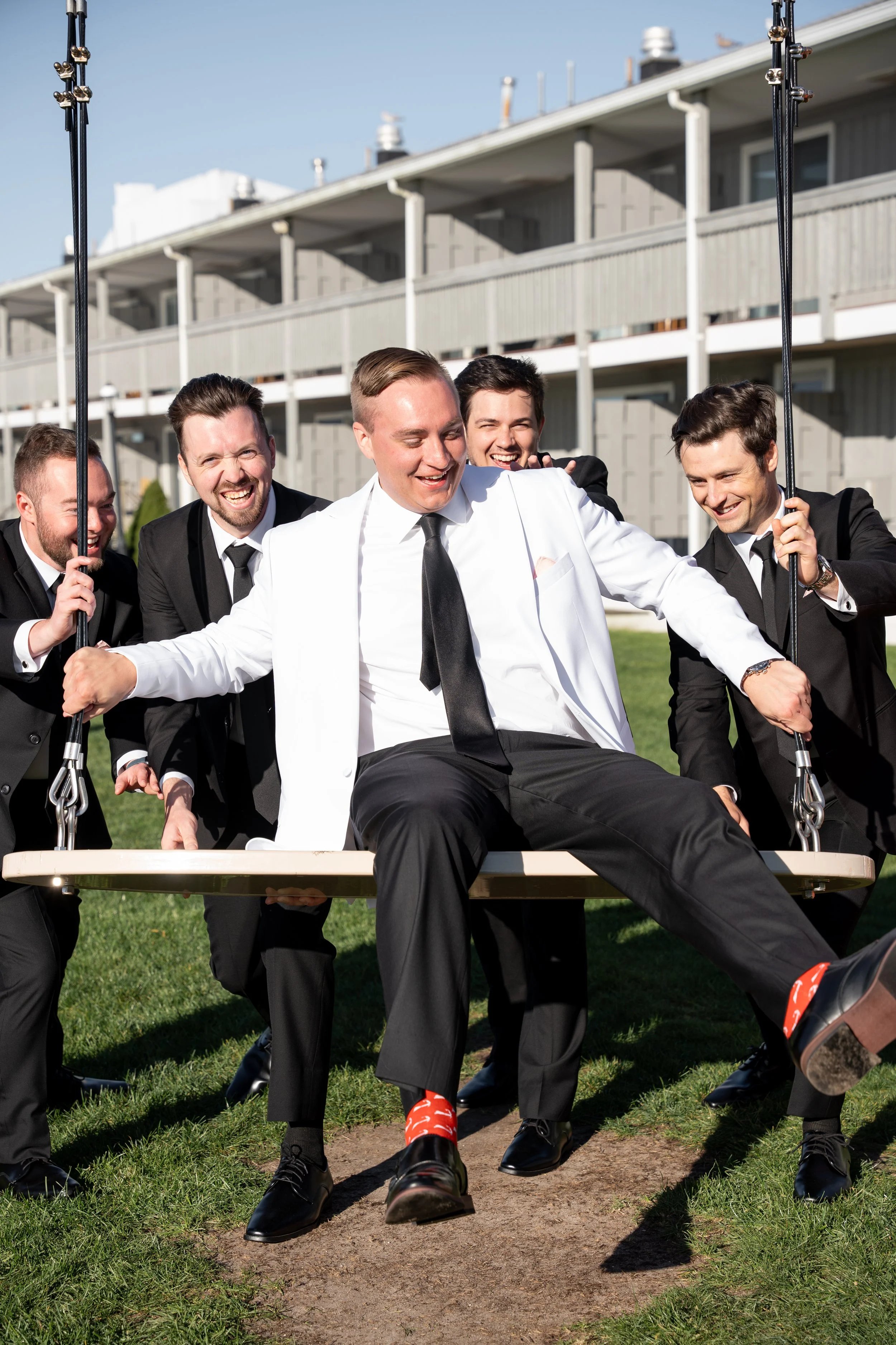 A group of five men dressed in suits, with one man in a white jacket, having fun on a swing on a grassy area outdoors, smiling and laughing, with a multi-story residential building in the background during a wedding at Red Jacket Resort in Cape Cod. 