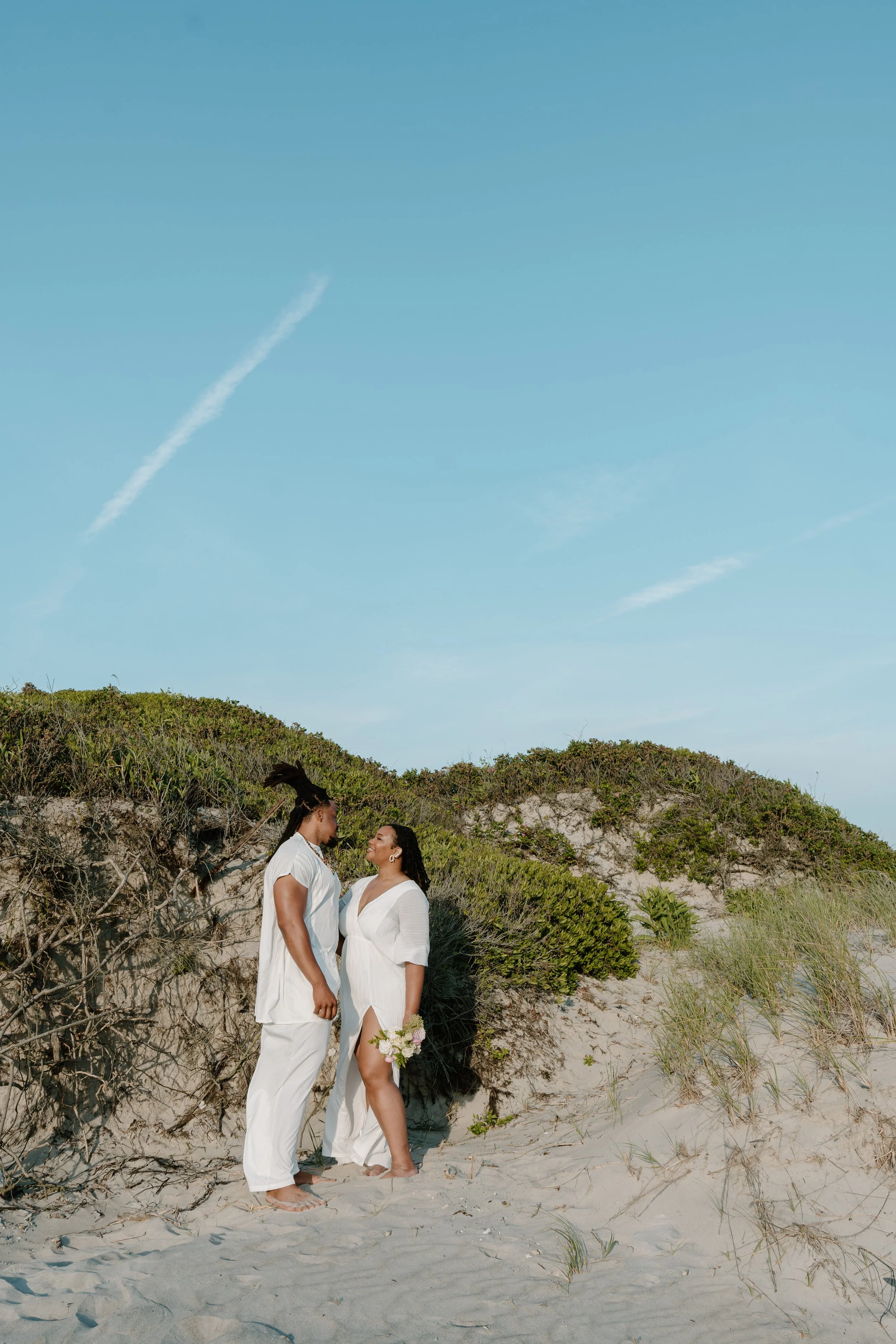 Couple dressed in white, standing on beach near dunes and green bushes, facing each other, holding flowers, under a blue sky at an elopement at Second Beach in Rhode Island. 