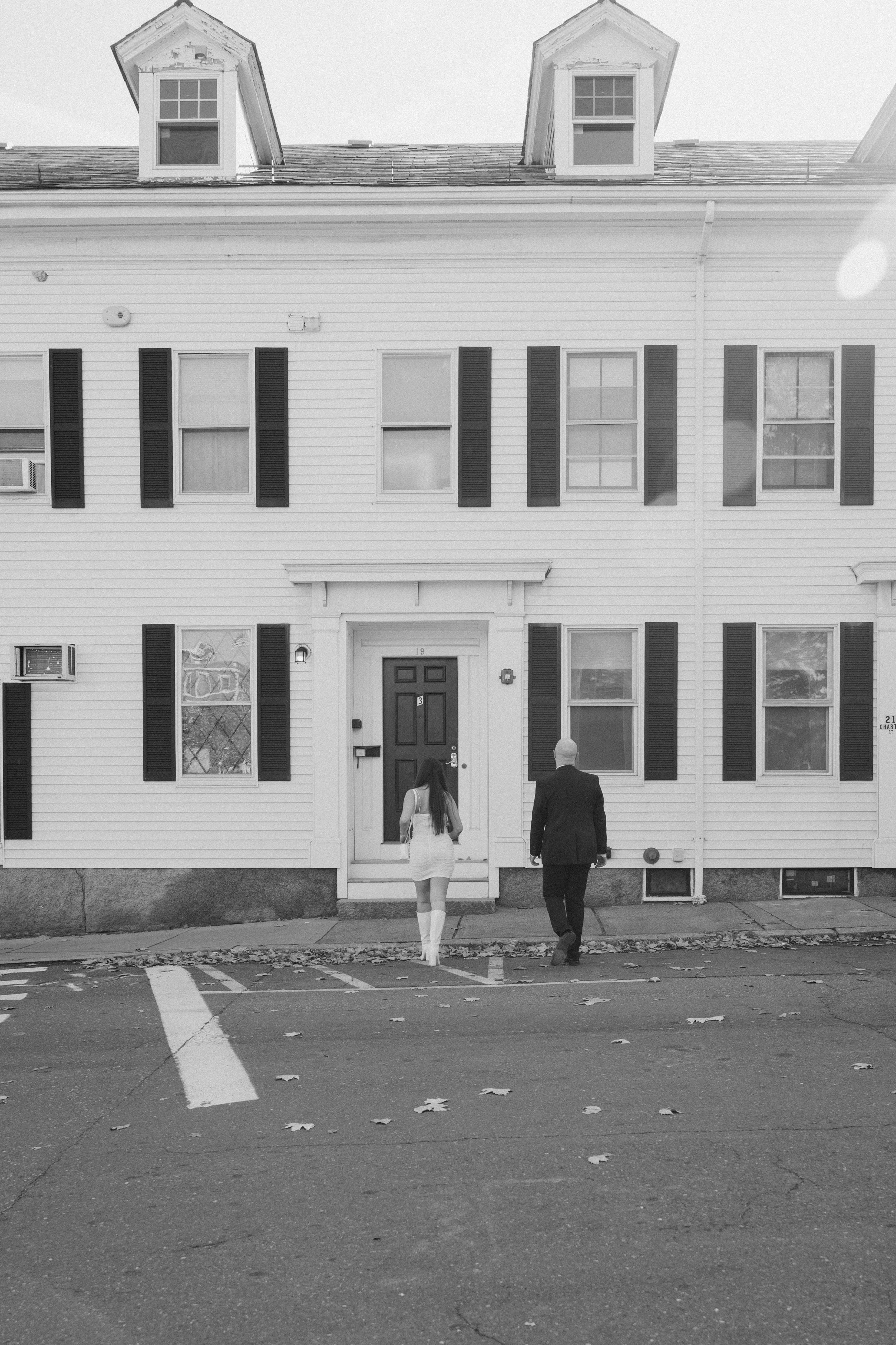 A black and white photo of a woman in a white dress and a man in a dark suit walking towards the entrance of a white house with black shutters on a residential street.