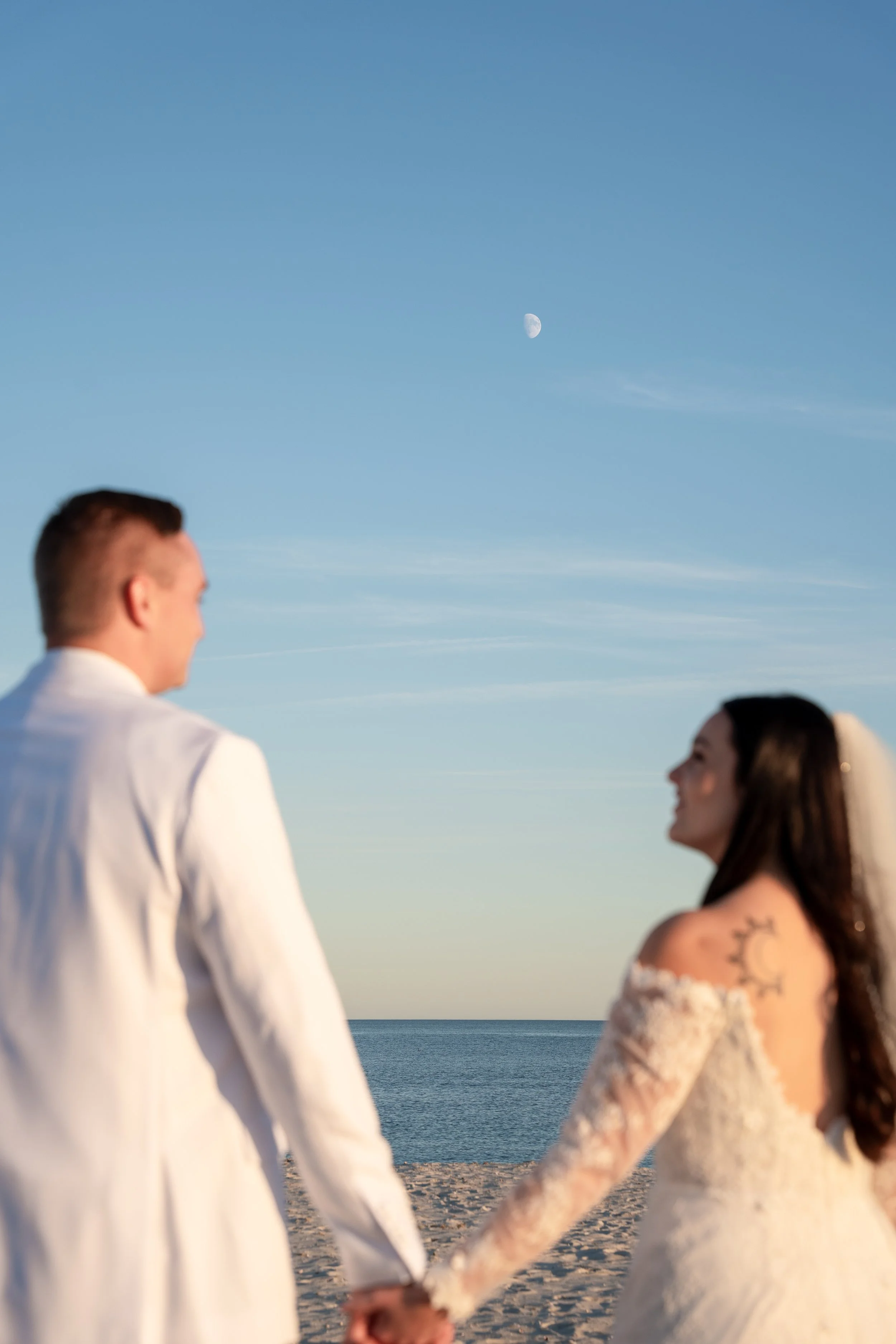A couple in wedding attire holding hands on a beach with the ocean and sky in the background, with the moon visible in the sky during a wedding at Red Jacket Resort in Cape Cod. 