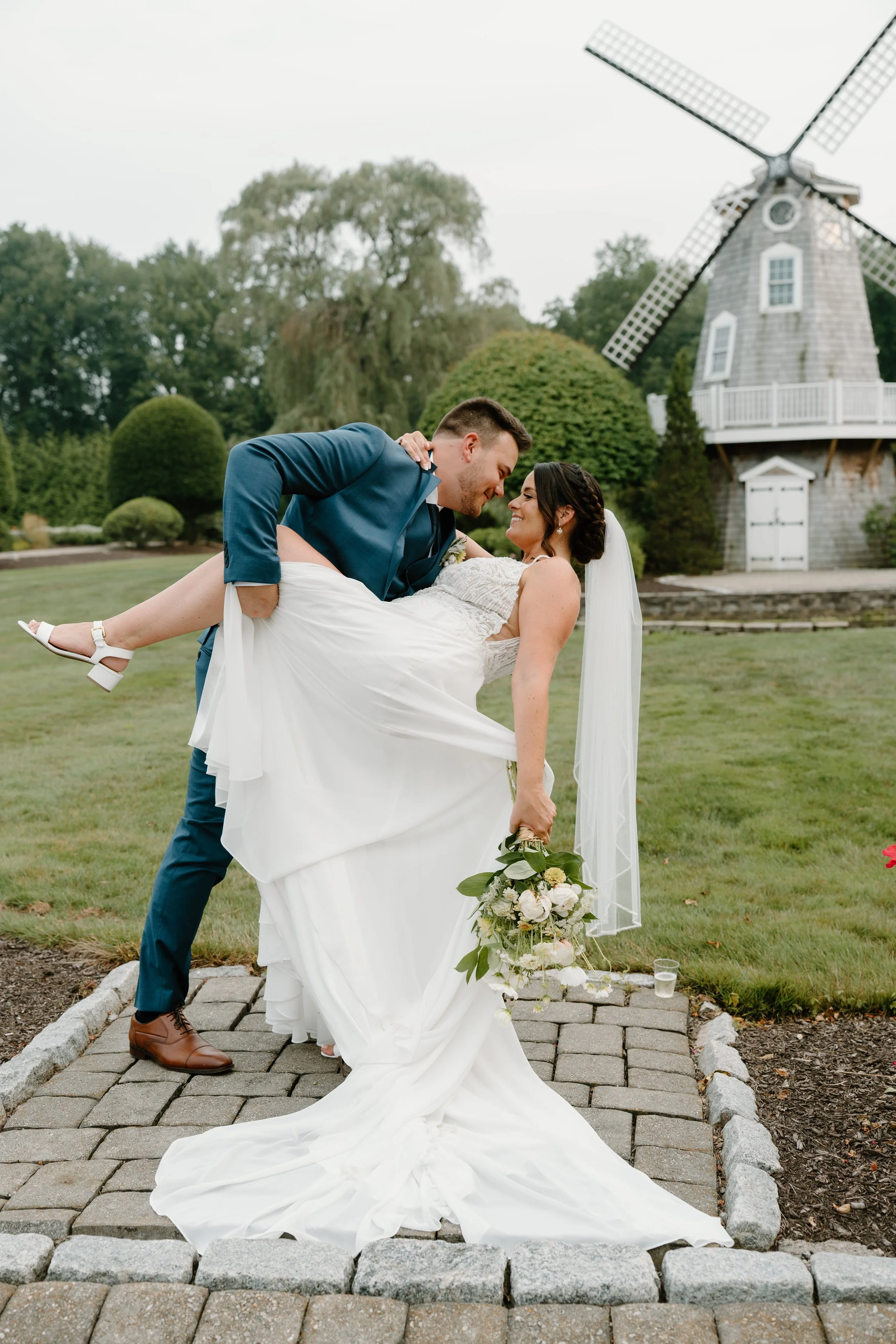 A newly married couple outdoors, with the groom lifting the bride, who is holding a bouquet, in front of a windmill and lush greenery during a wedding at the Aqua Turf Club in Connecticut. 