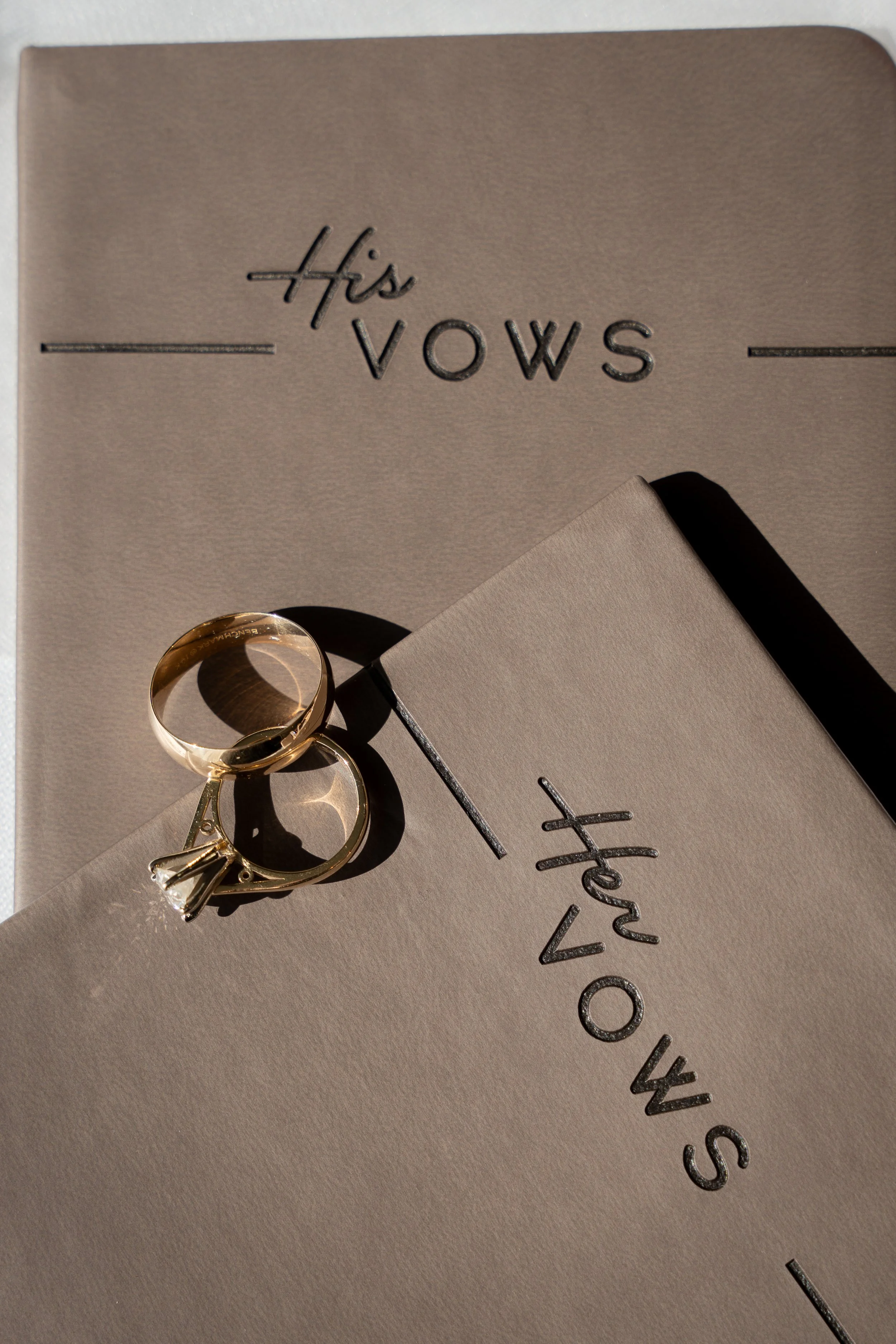 Close-up of wedding rings resting on a beige VOWS book and a matching card with the words "His" and "Her Vows" embossed on them, with sunlight casting shadows during a wedding at Red Jacket Resort in Cape Cod. 