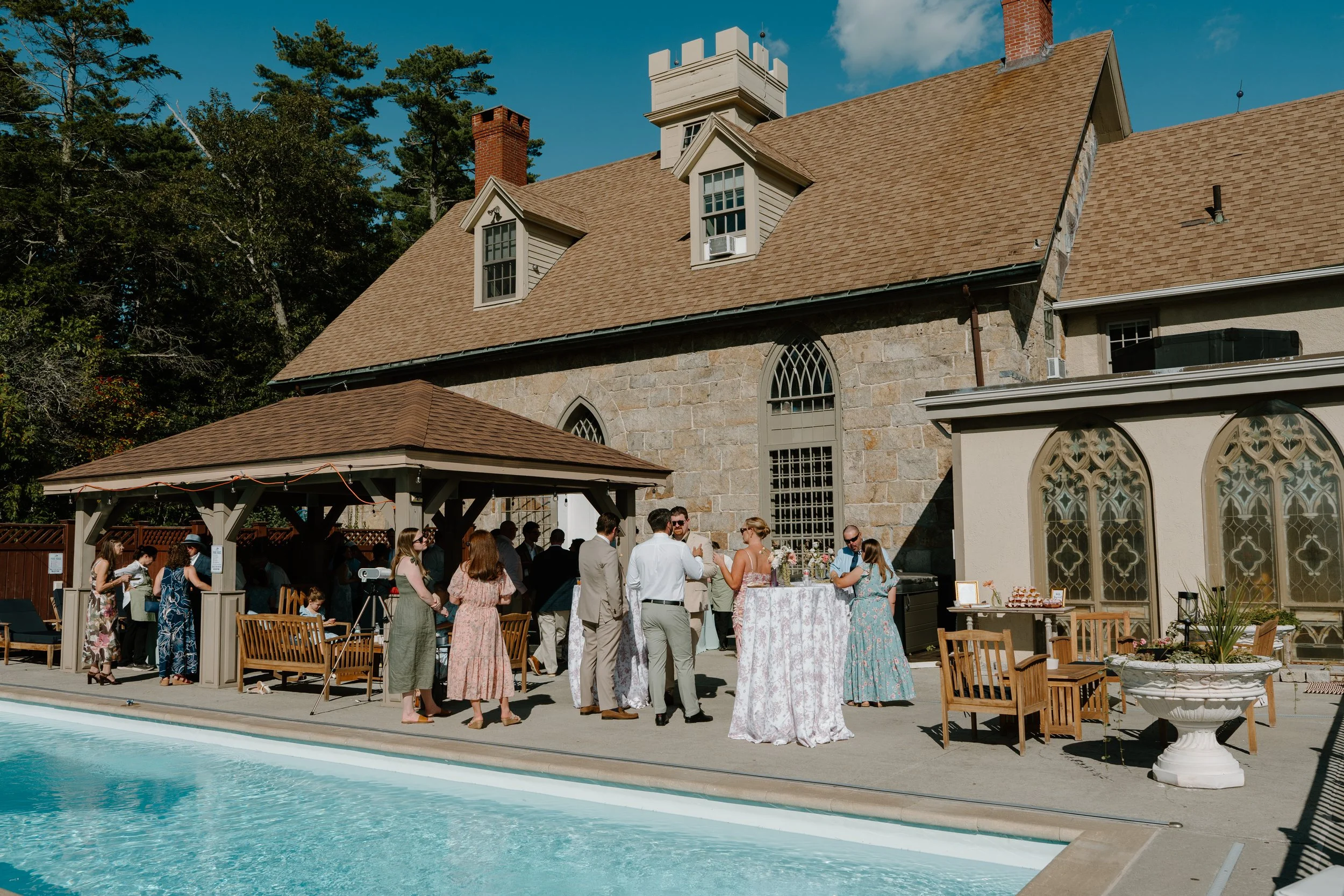 People gathered outdoors near a pool during a social event at a large stone and stucco house with a tan shingle roof and large gothic-style windows, under a blue sky with a few clouds at a wedding in Bath, Maine. 
