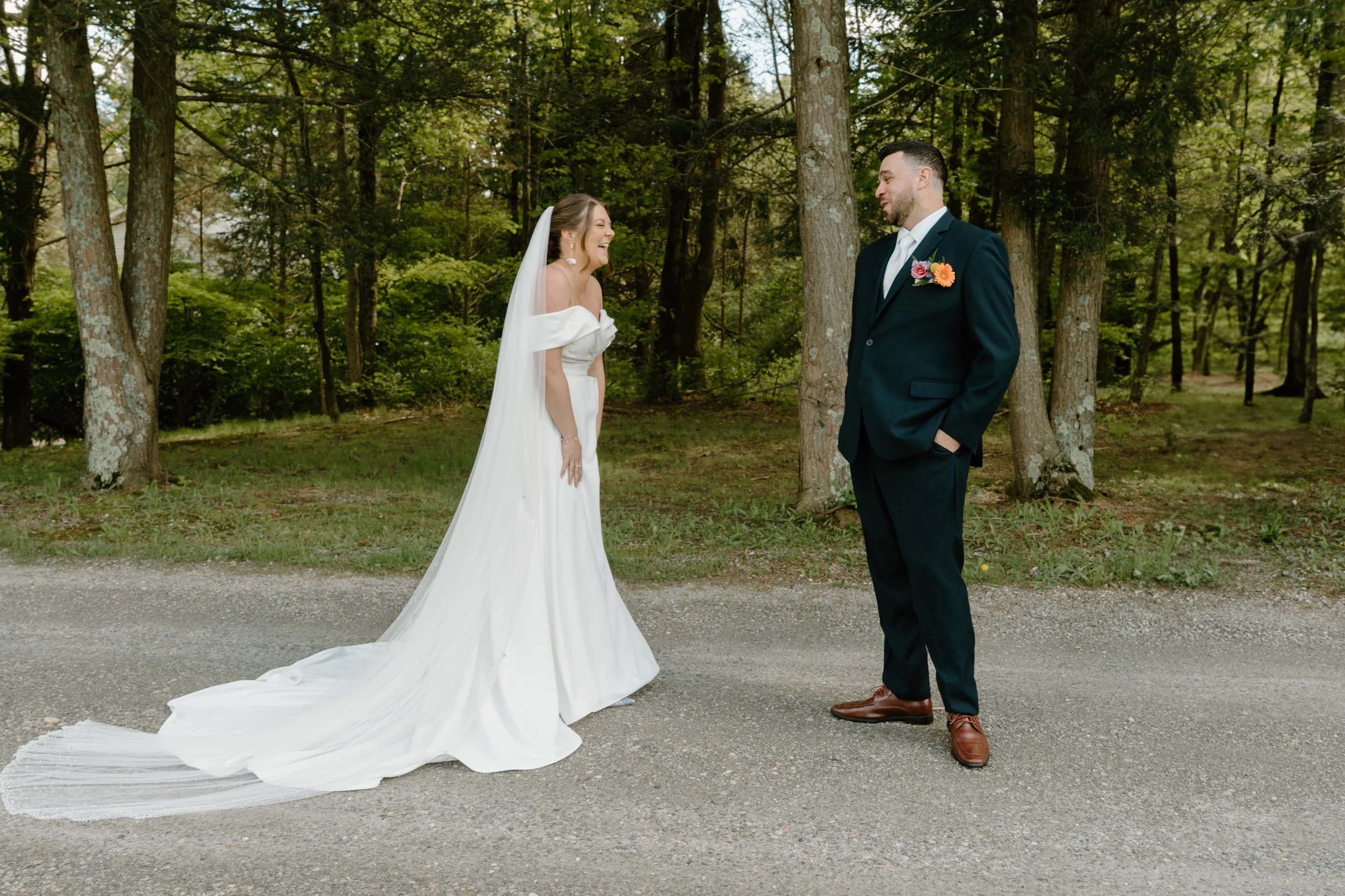 A bride in a white wedding dress with a train standing on a gravel path, smiling and laughing at a groom in a navy suit for during their first look at a wedding at Holiday Hill Day Camp in Connecticut.