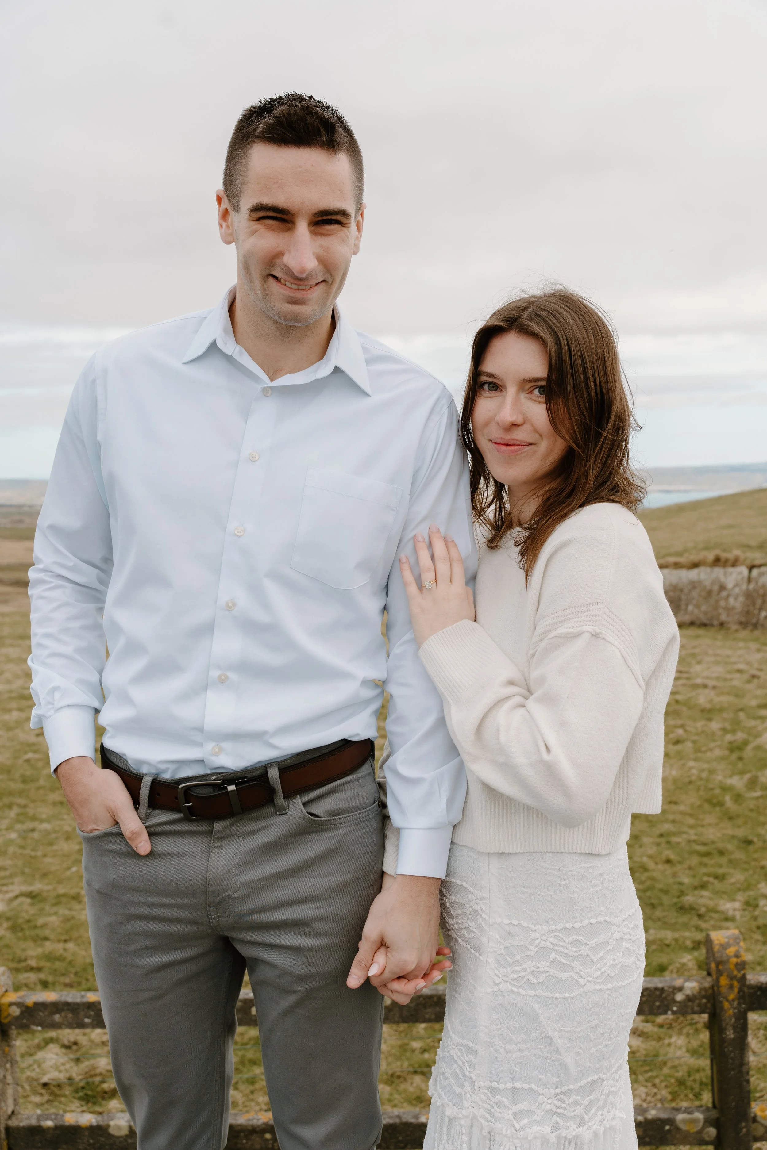 A happy couple holding hands outdoors in a rural landscape with rolling hills and cloudy sky for an engagement photo session in Galway, Ireland. 