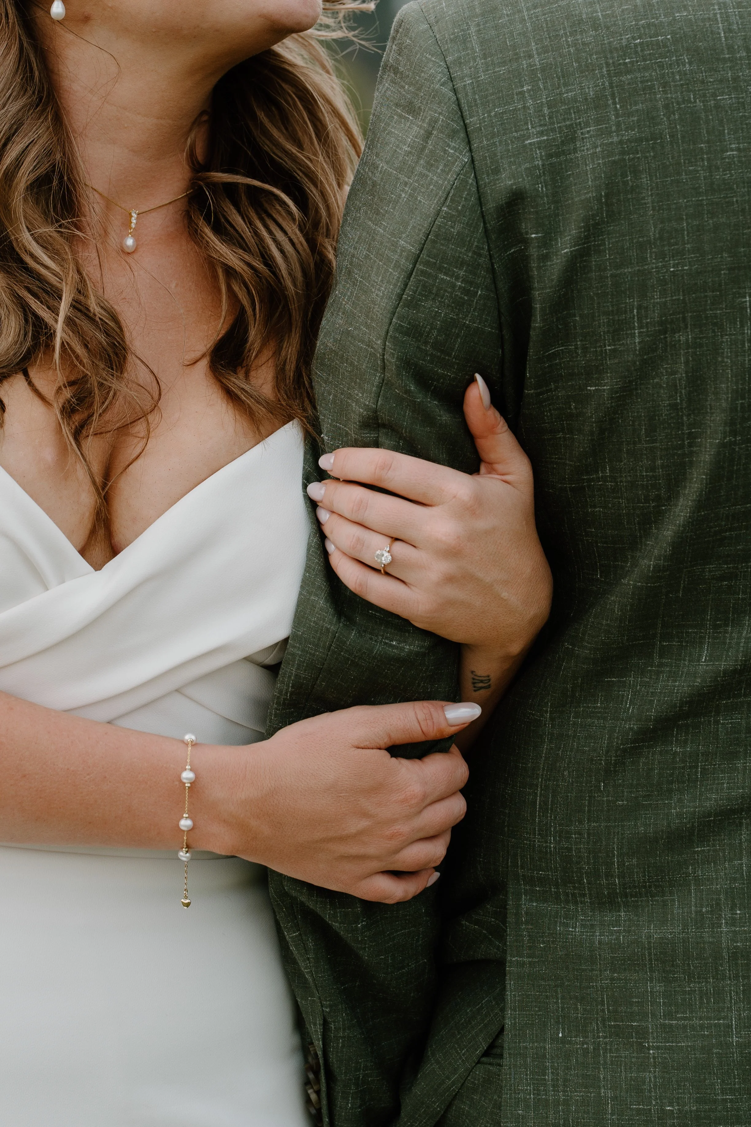 A woman in a white dress with pearl jewelry holds onto a man in a green suit jacket, showing her wedding ring and a tattoo on her wrist at a wedding at Everett Ranch in Salida, Colorado.