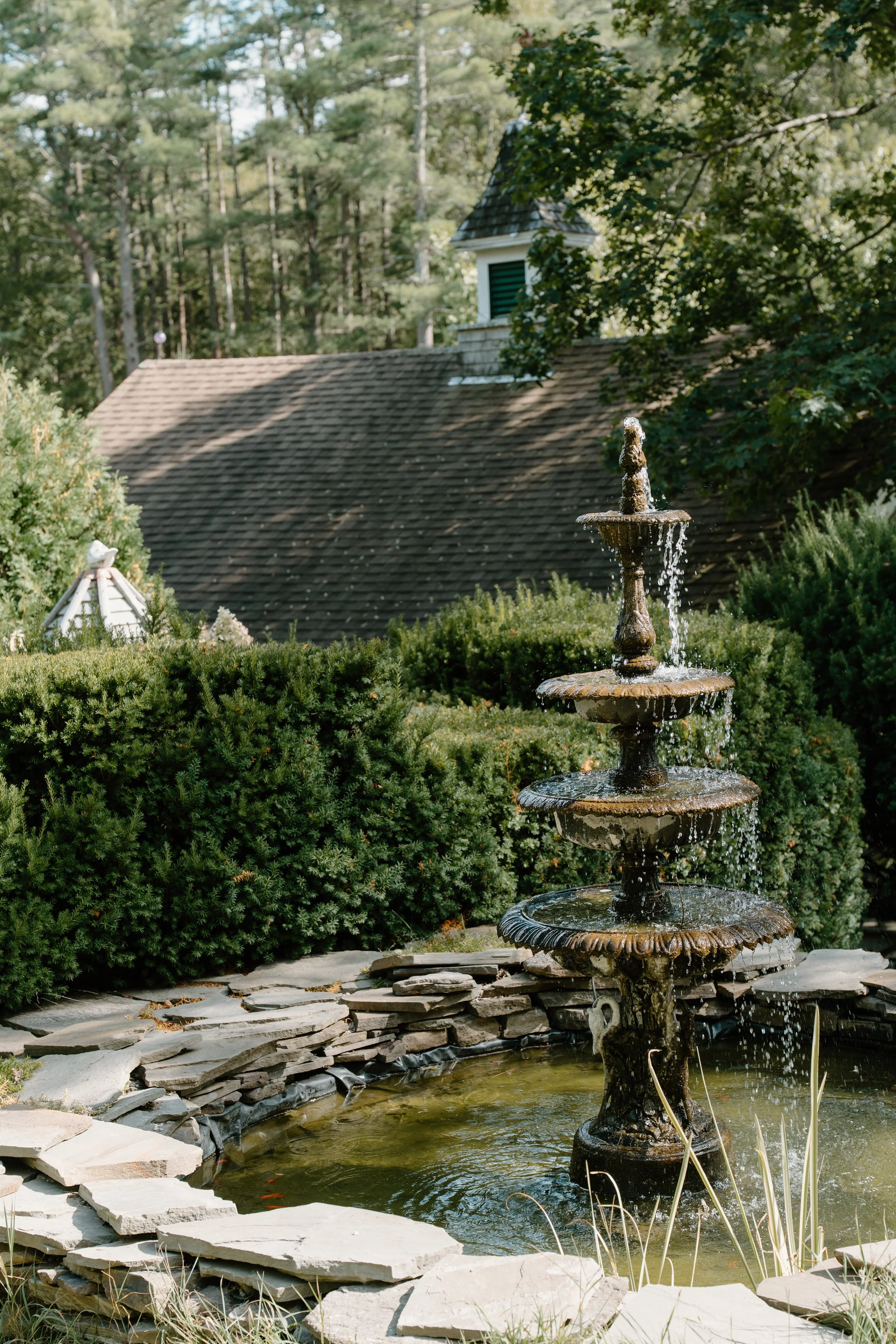 A multi-tiered stone fountain in a garden with green bushes and trees, a pond at the base, and a house with a steep roof and small turret in the background at a wedding in Bath, Maine. 