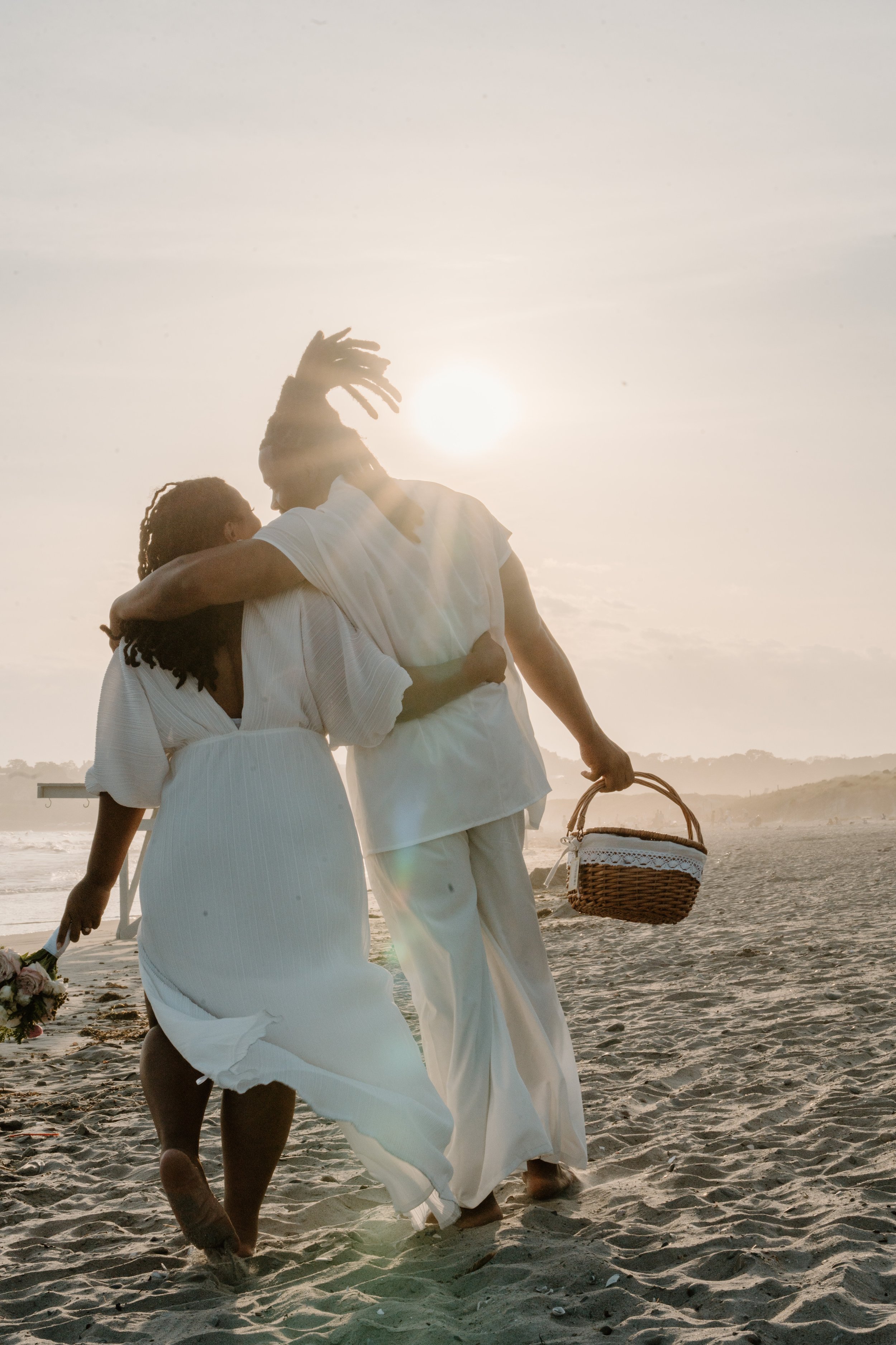 A couple walking on the beach at sunset, dressed in white, with the woman holding a bouquet and the man carrying a picnic basket at an elopement at Second Beach in Rhode Island. 