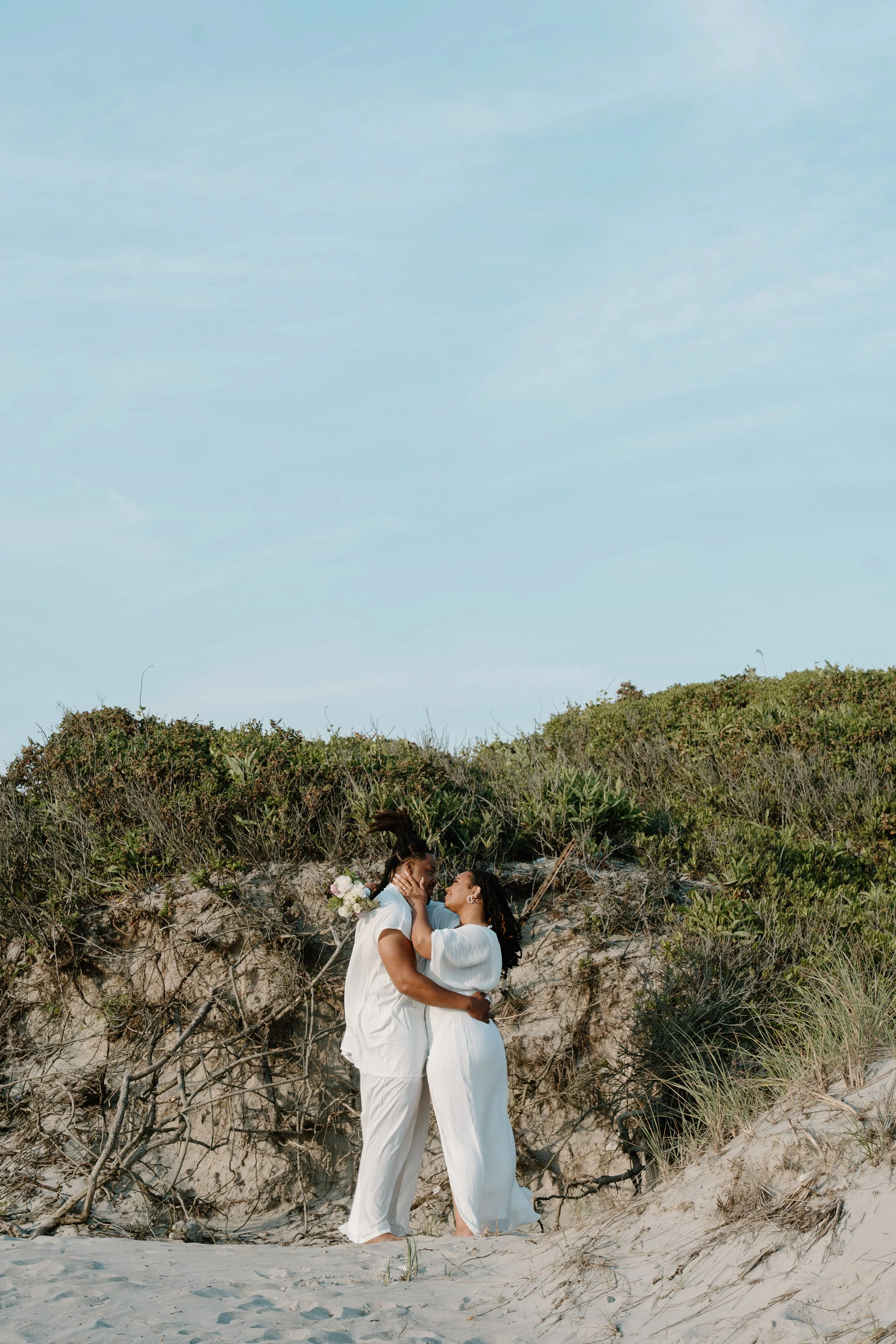 A couple dressed in white holding each other on a sandy beach with bushes behind them, embracing and about to kiss at an elopement at Second Beach in Rhode Island. 