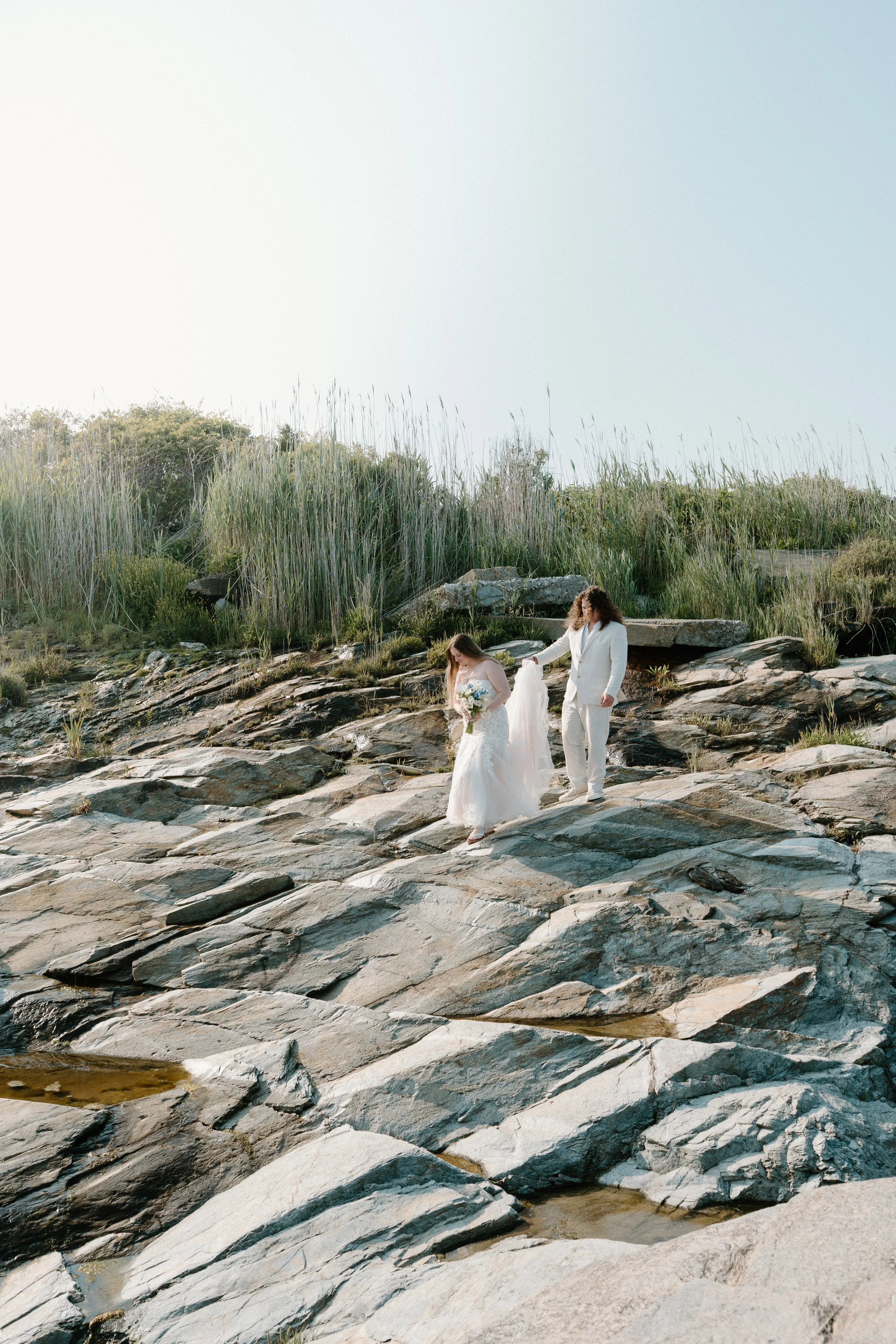 A couple dressed in white wedding attire walking on rocky terrain near a body of water, holding hands with lush green plants in the background at an elopement at Beavertail State Park in Rhode Island. 