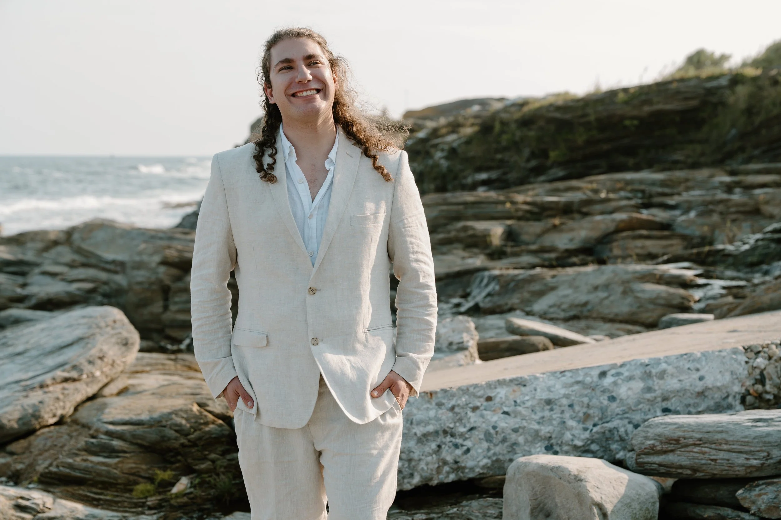 A young man with long curly hair wearing a beige suit standing on rocky shoreline near the ocean, smiling at the camera at an elopement at Beavertail State Park in Rhode Island. 