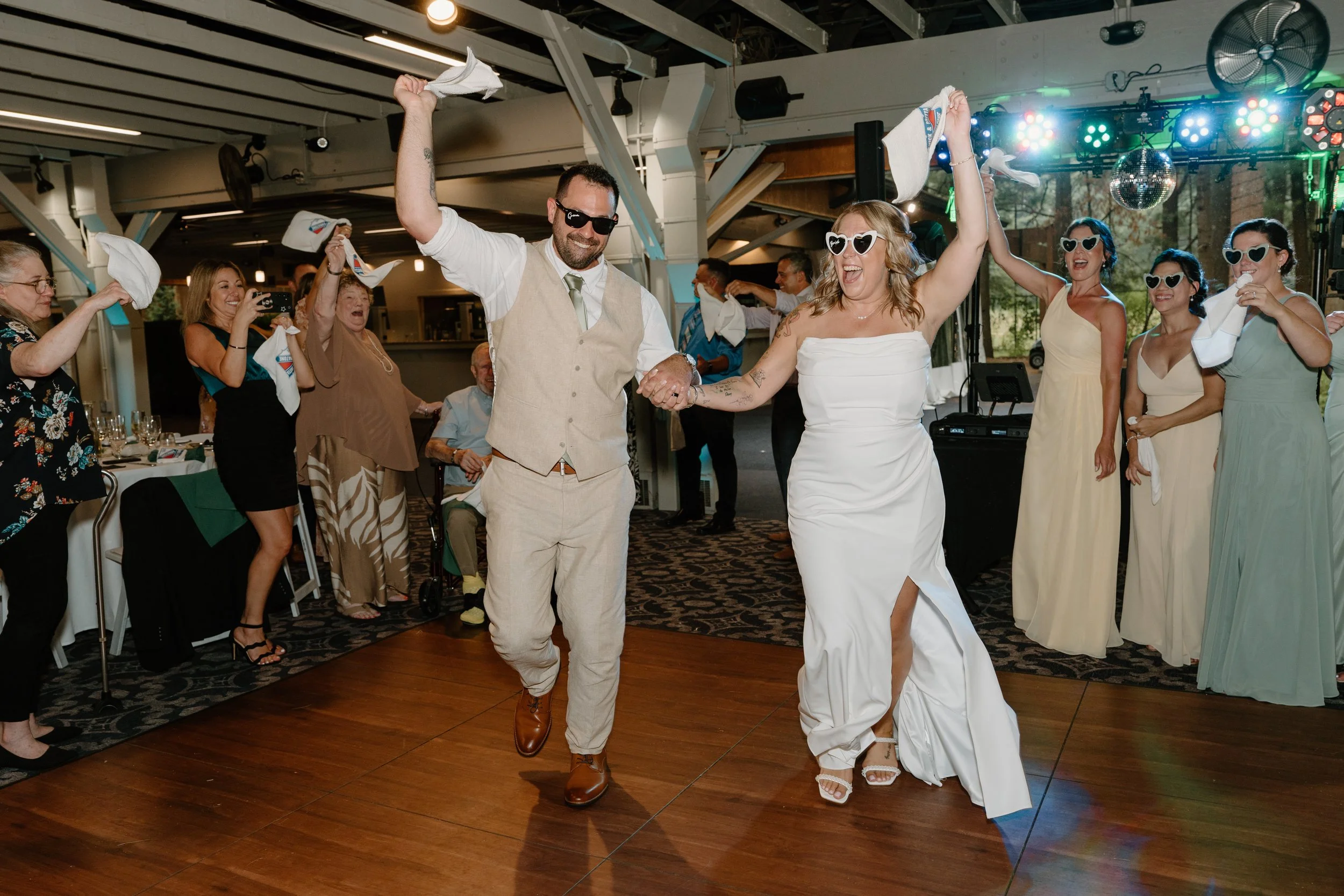 Bride and groom dancing at wedding reception, surrounded by guests, some wearing heart-shaped sunglasses, with a band playing in the background for a wedding at Tunxis Country Club in Connecticut. 