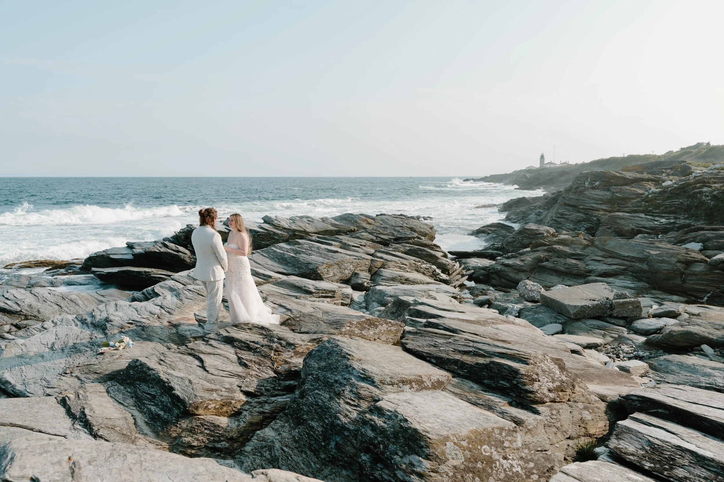A couple in wedding attire exchanging vows on a rocky beach with the ocean and a lighthouse in the background at an elopement at Beavertail State Park in Rhode Island. 