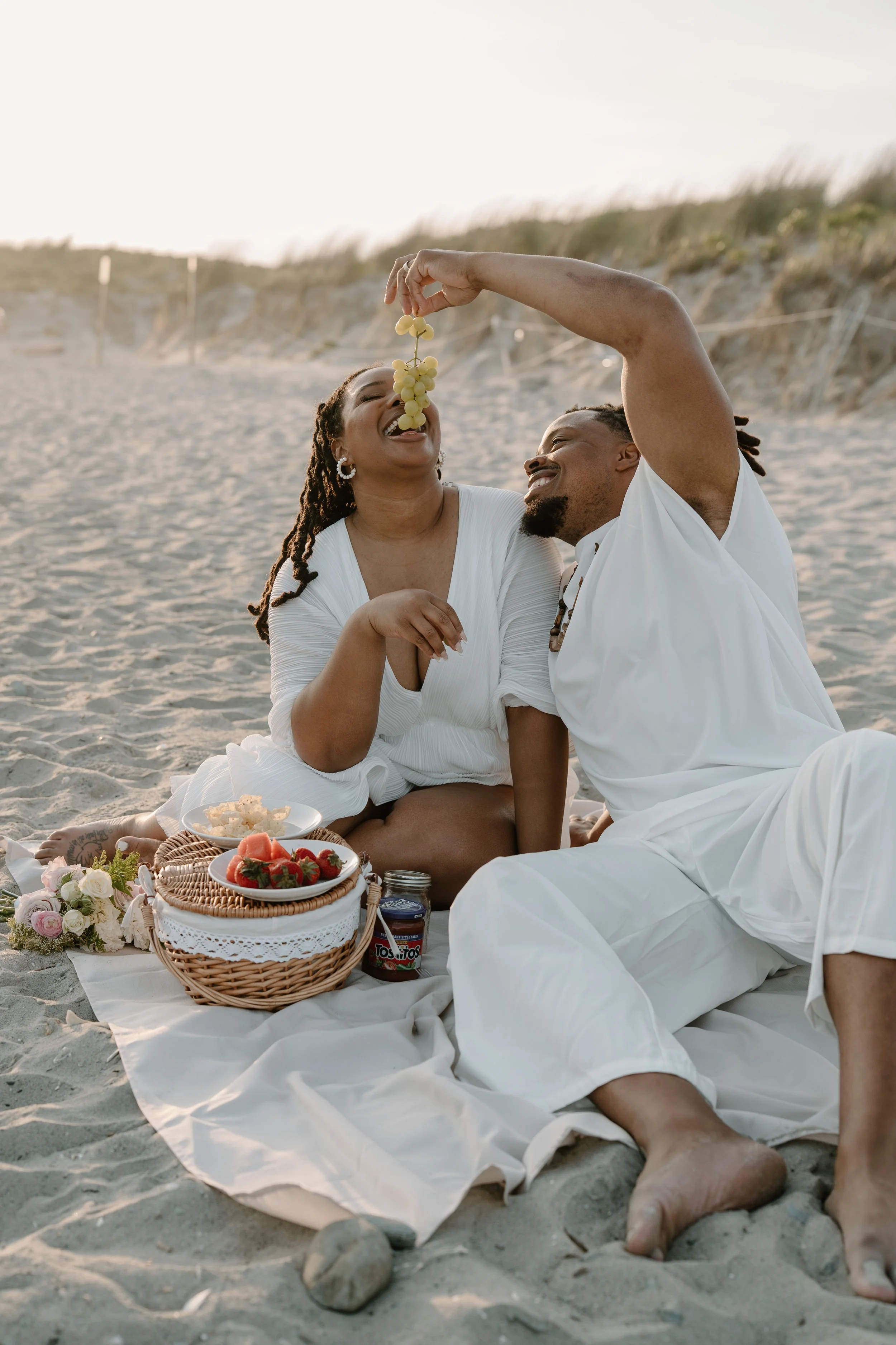 A happy couple having a picnic on the beach, with the man holding a bunch of grapes over the woman's mouth, both smiling and enjoying their time together at an elopement at Second Beach in Rhode Island. 