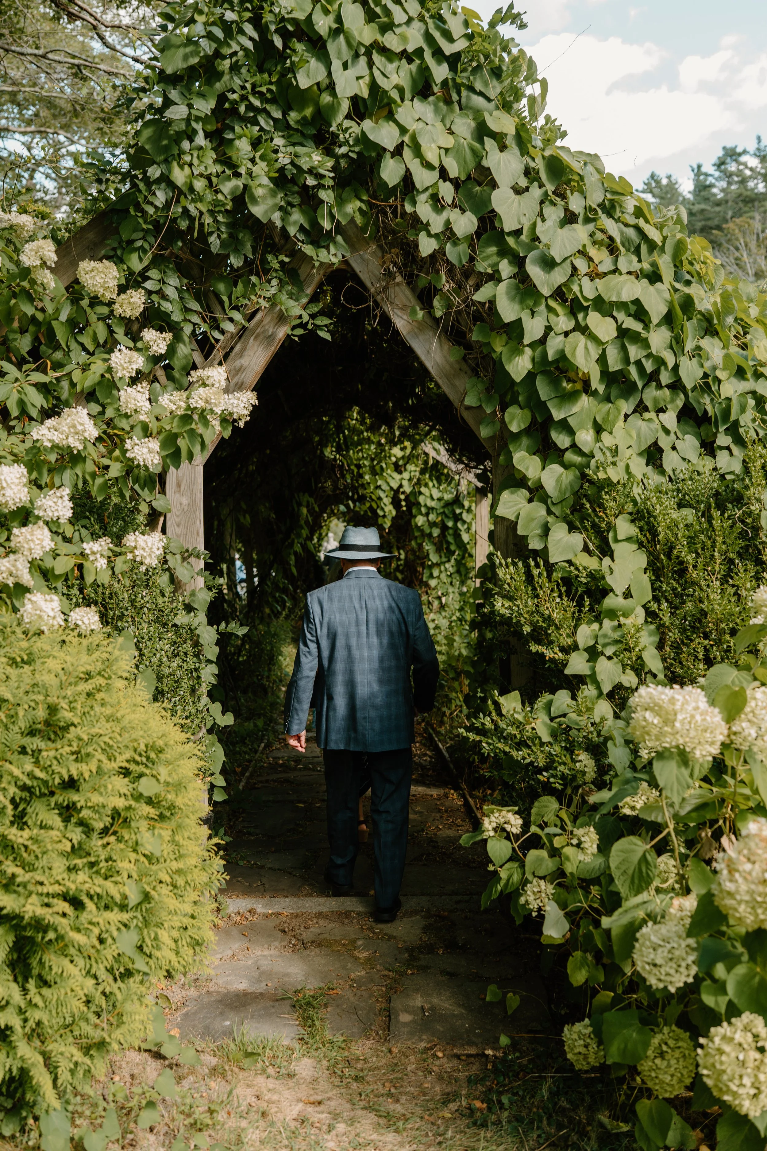 A man in a suit and hat walking through a garden archway covered in green foliage and white flowers at a wedding in Bath, Maine. 